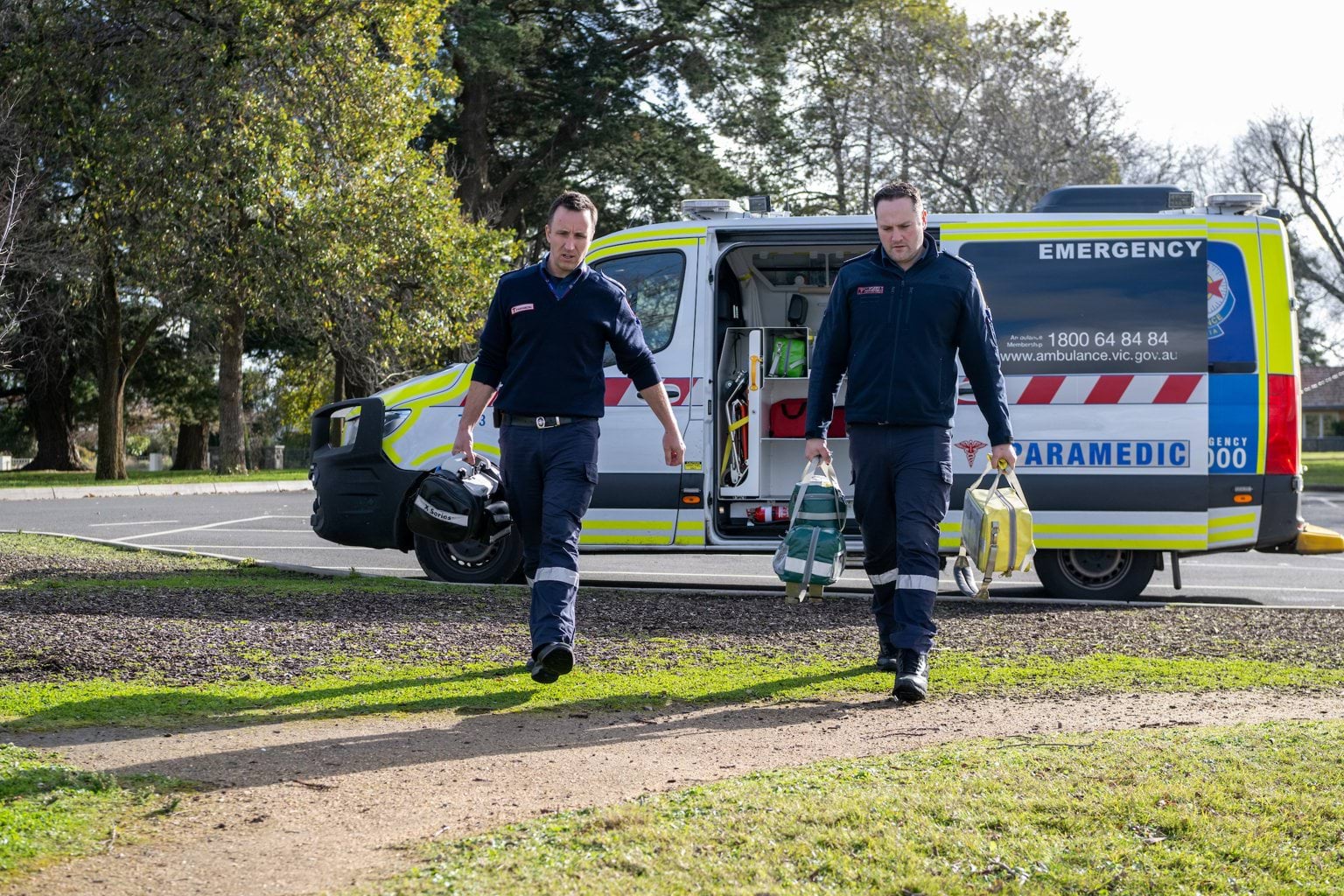 Two male paramedics walk out of the ambulance carrying equipment bags.