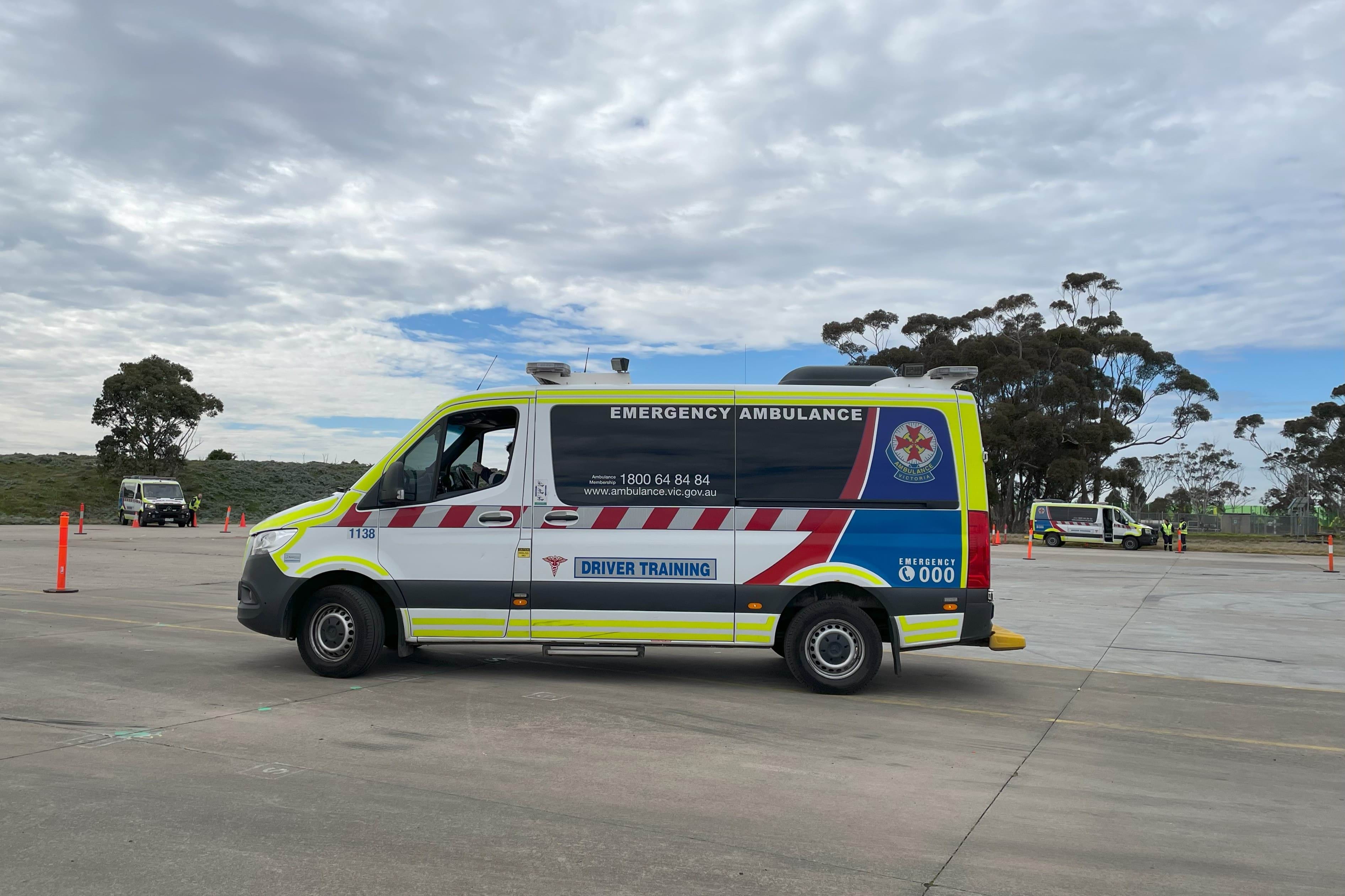 An Ambulance Victoria Driver Training ambulance. Two more ambulances are in the background. There are orange cones to drive around.