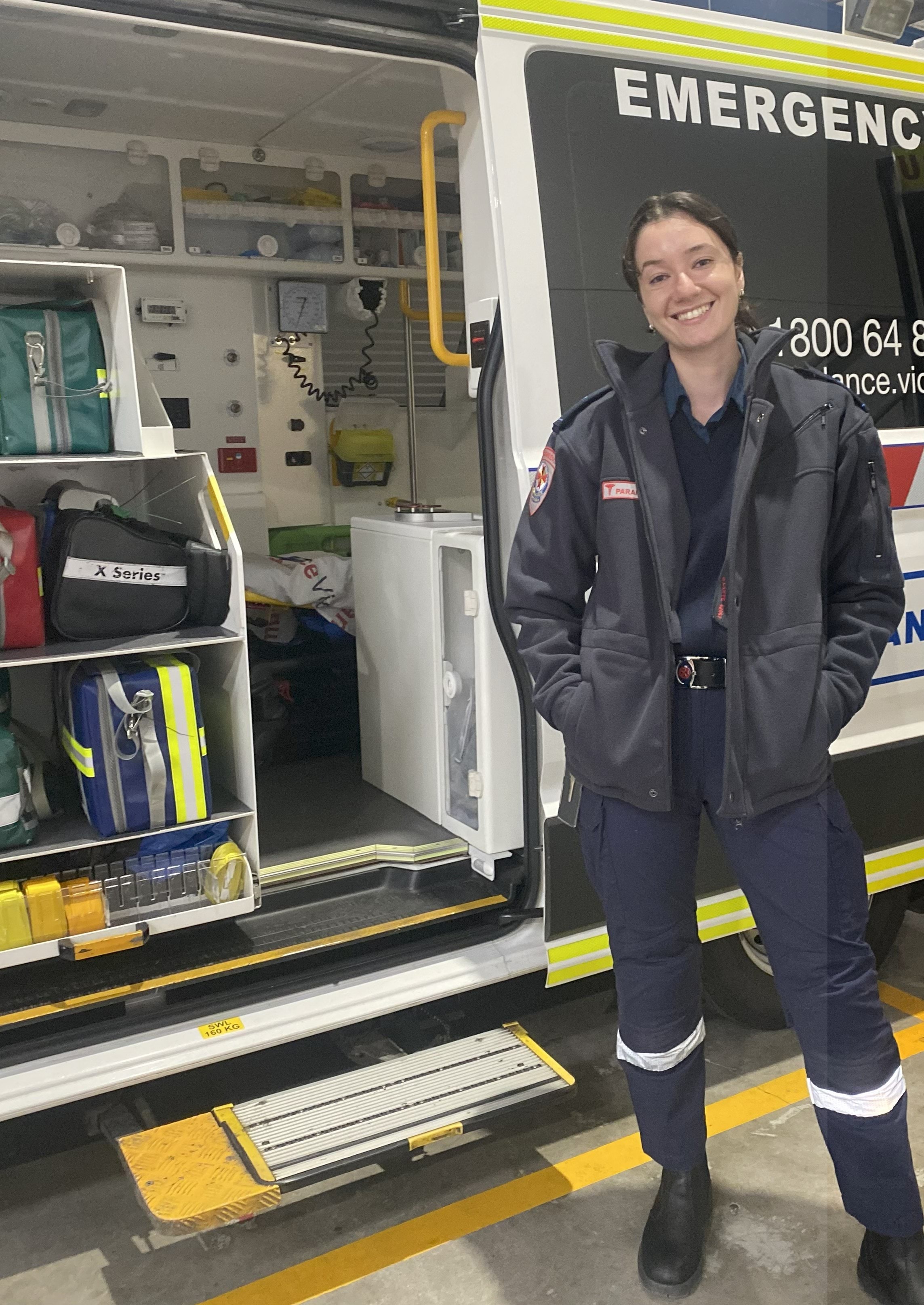 A female paramedic smiles for the camera in front of the open side door of an ambulance.