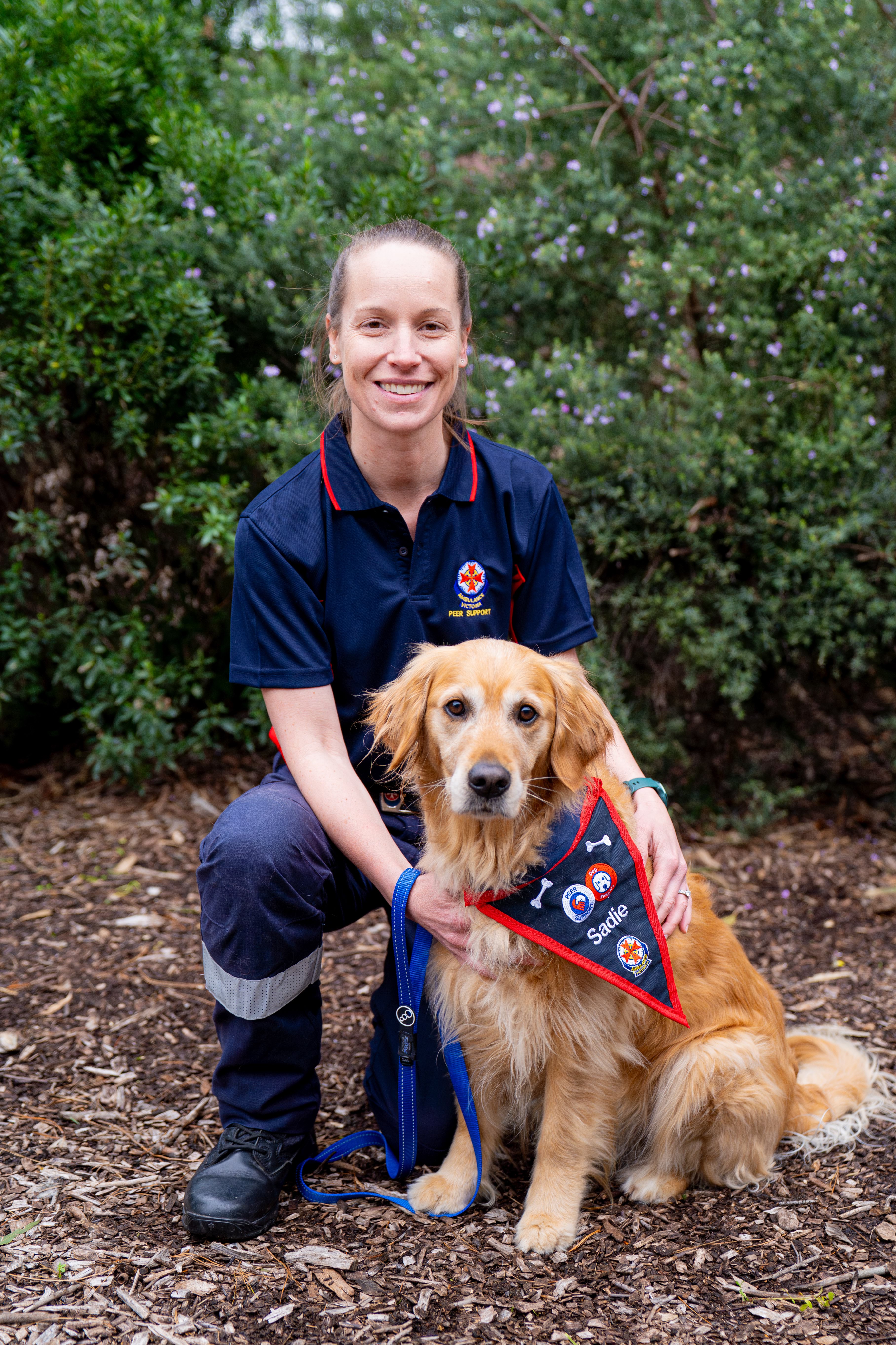 A woman in Ambulance Victoria uniform crouches next to her dog, who wears an AV bandana that says 'Sadie'.