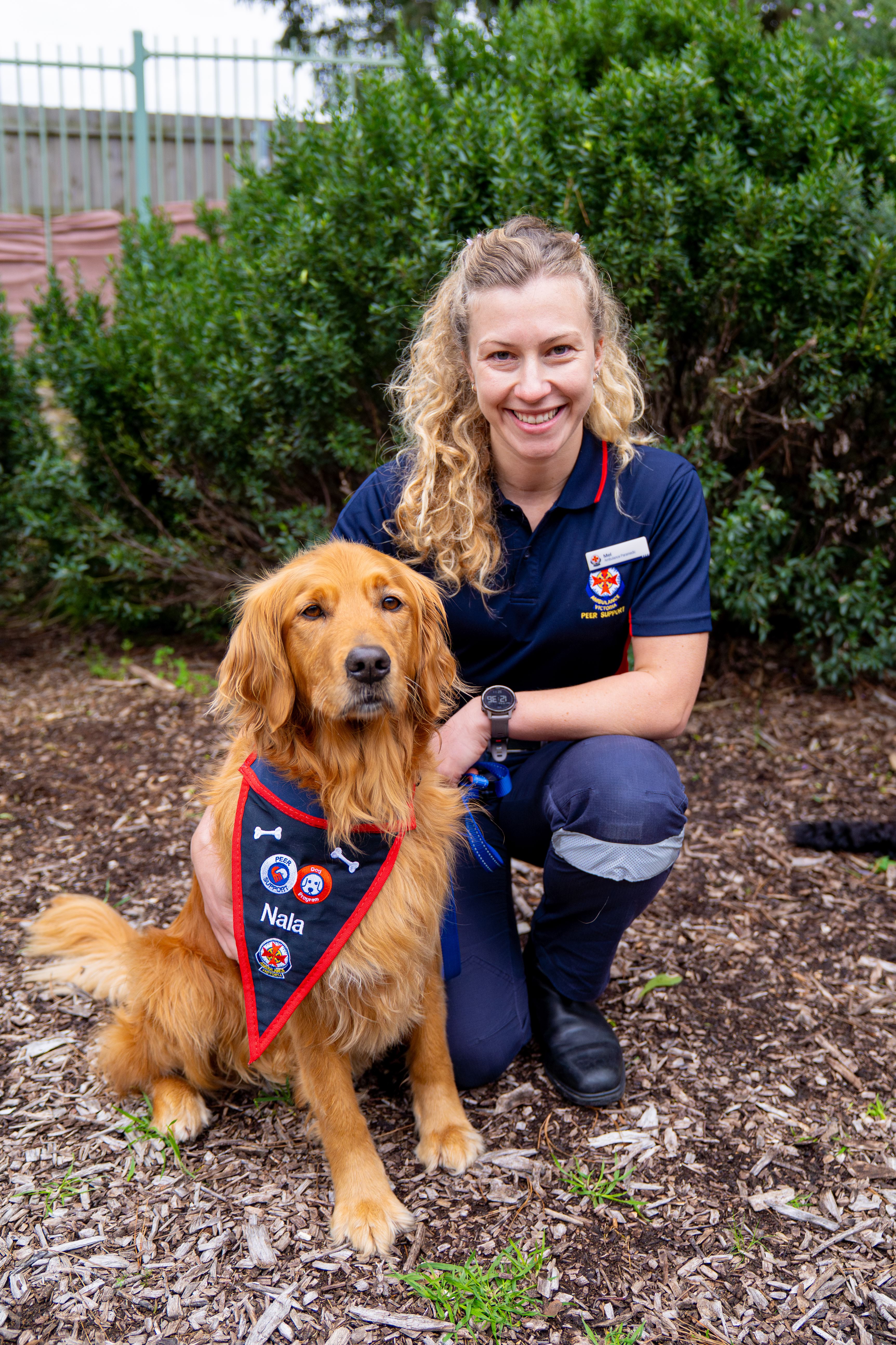 A woman in Ambulance Victoria uniform crouches next to her dog, who wears an AV bandana that says 'Nala'.