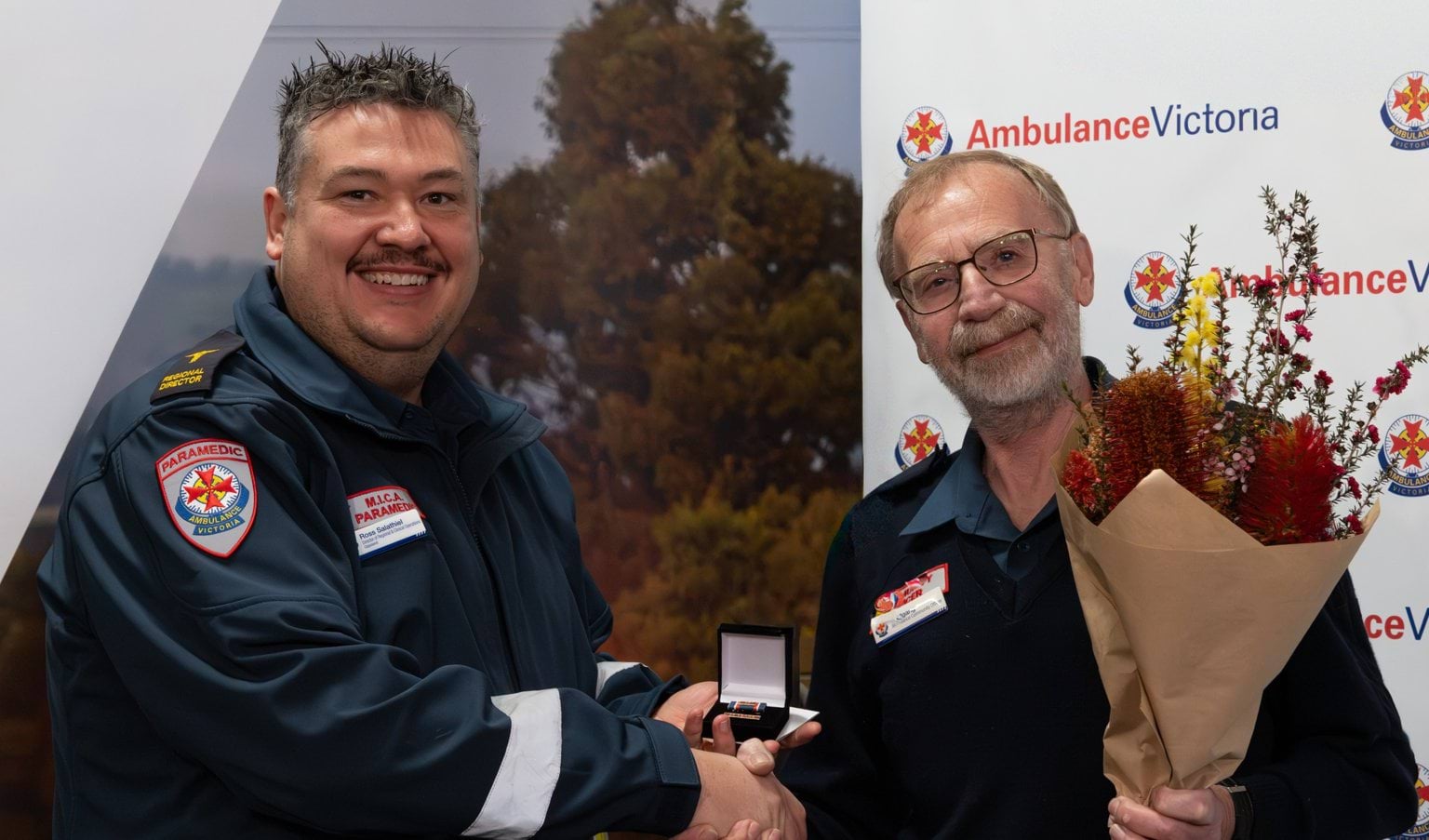 A man in AV uniform presents another man in AV uniform with a medal and flowers.