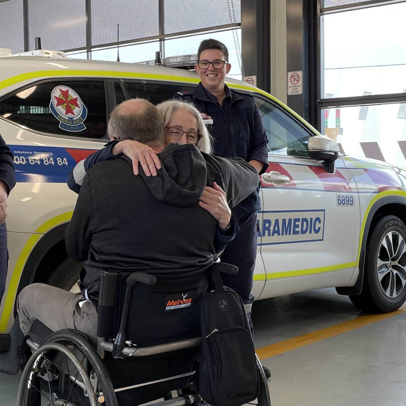 A paramedic hugs a man in a wheelchair. Another paramedic smiles in the background.