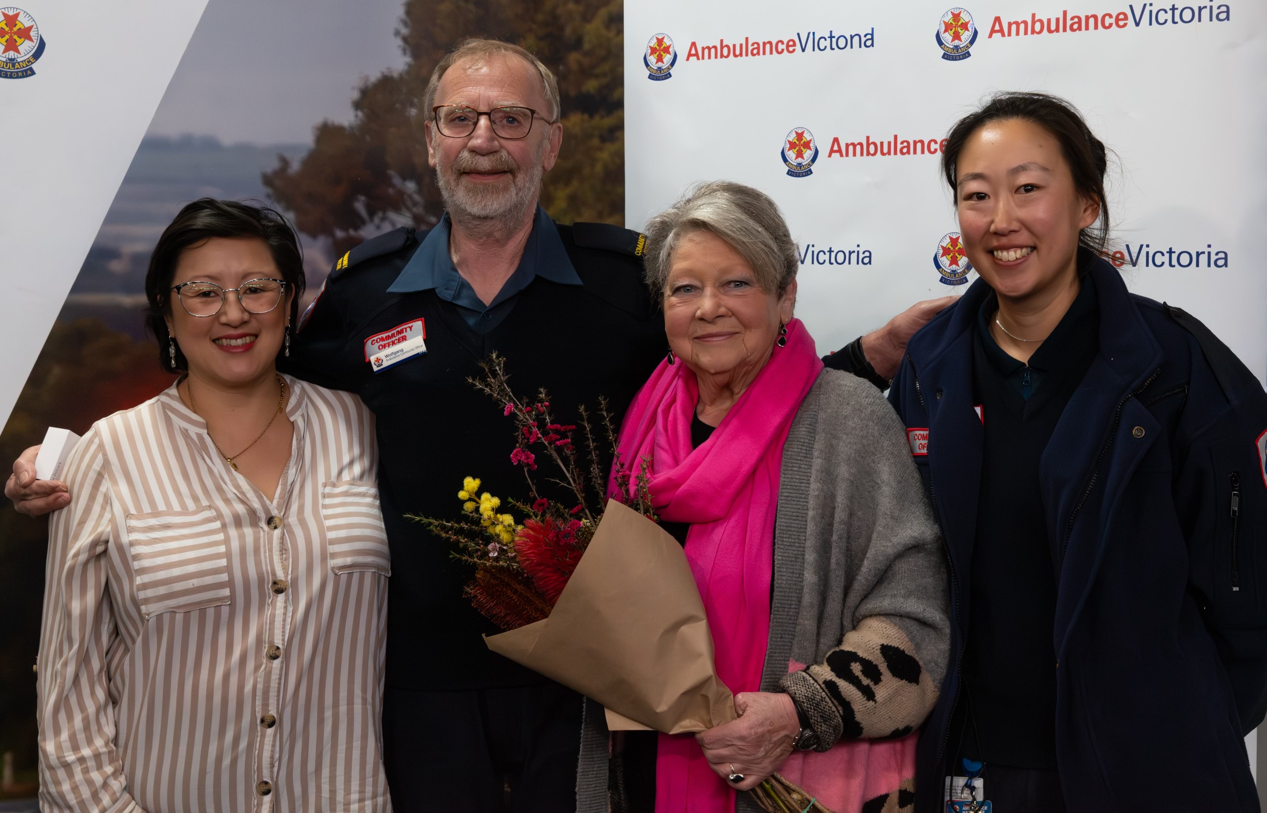A man in Ambulance Victoria uniform smiles with his arms around three women. The woman on the right is also in Ambulance Victoria uniform.