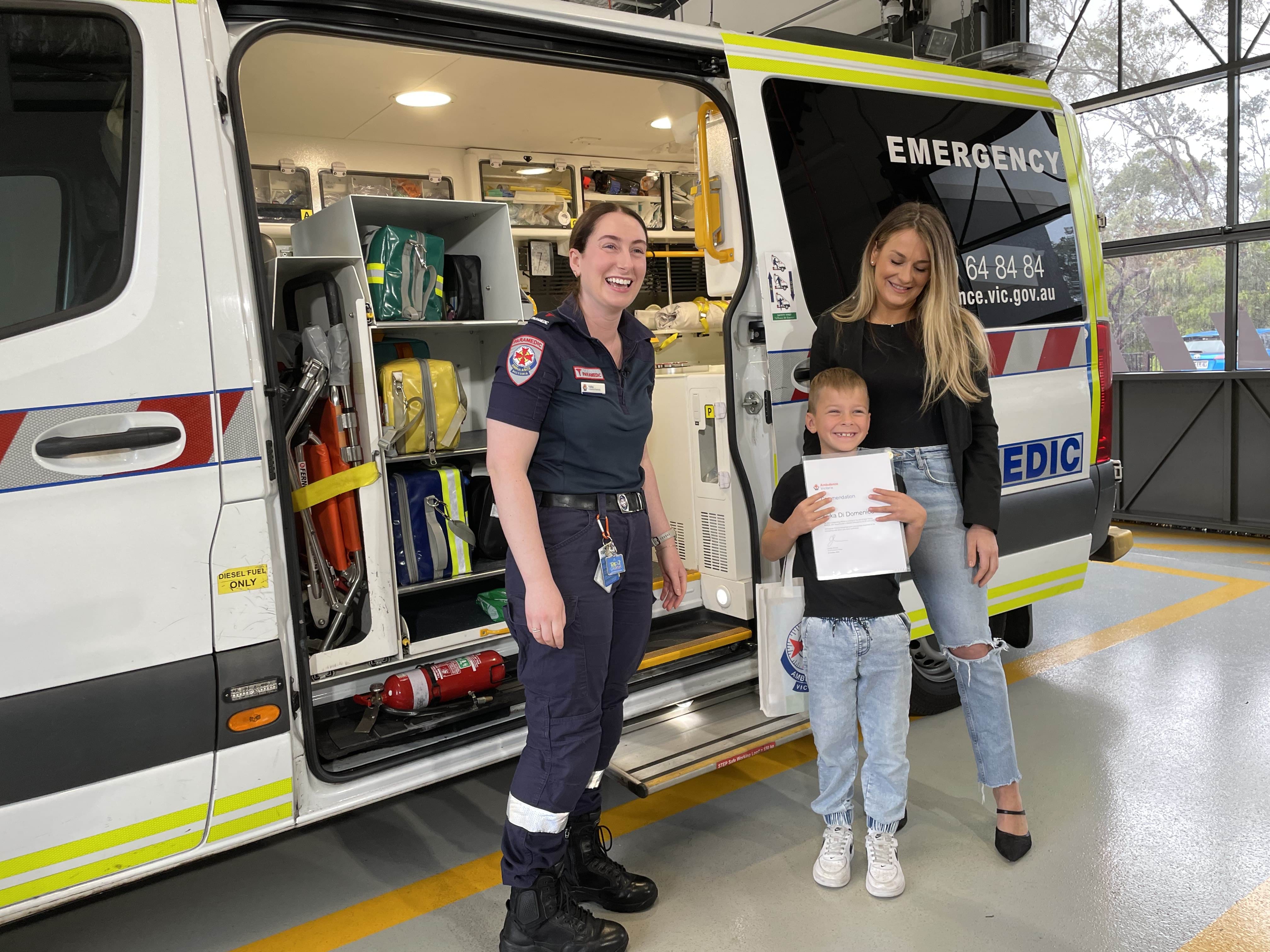 A boy grins holding a certificate. He stands next to his mum and a paramedic, in front of an ambulance.