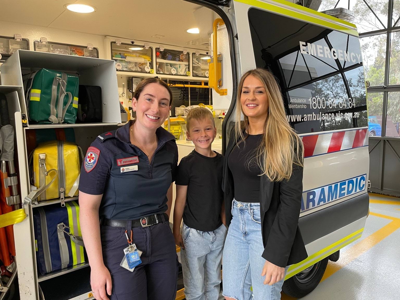 A paramedic smiles for the camera alongside a young boy and a woman. They stand at the side door of an ambulance.