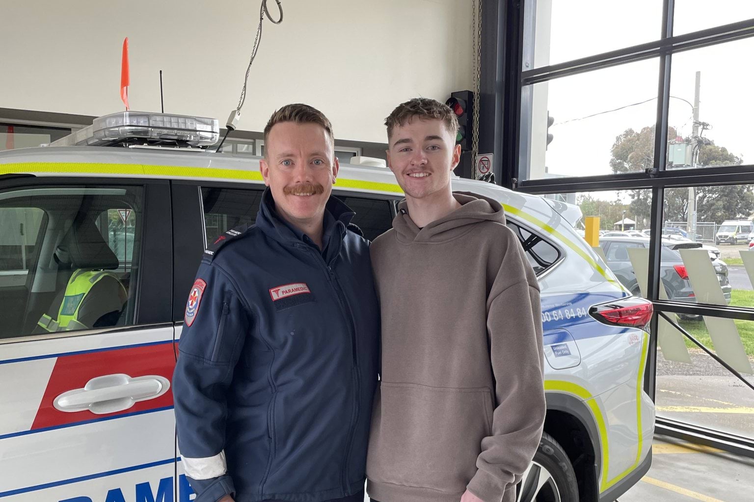 A paramedic and a young man in casual clothes smile for the camera, in front of an Ambulance Victoria car.