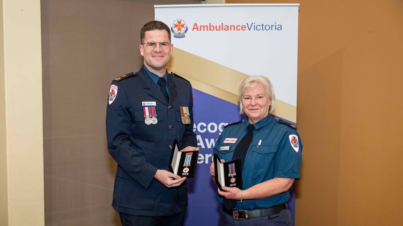 A man and woman in formal Ambulance Victoria uniform, holding award medals.