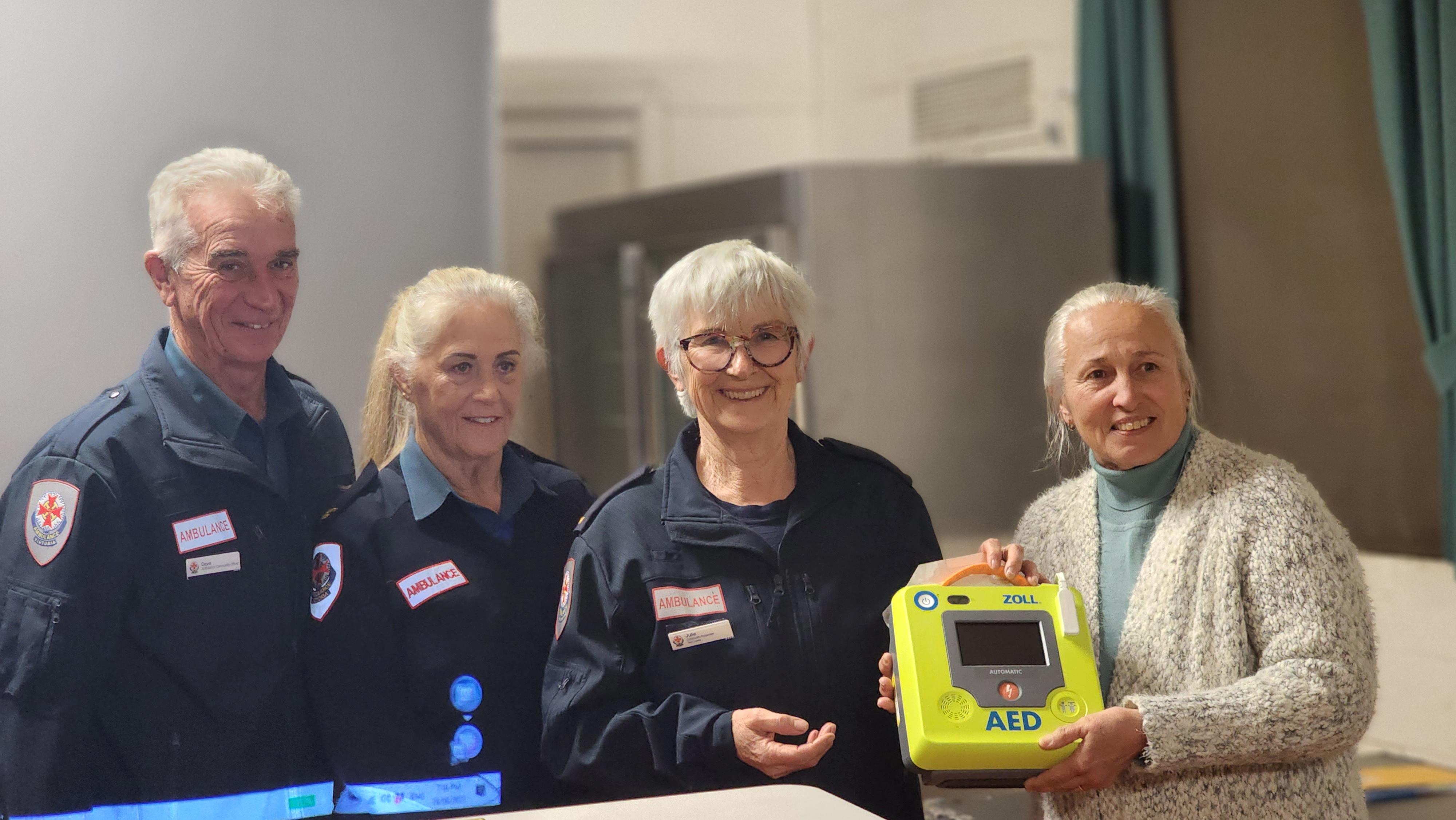 A group of three smiling first responders in uniform with another woman holding a defibrillator. 