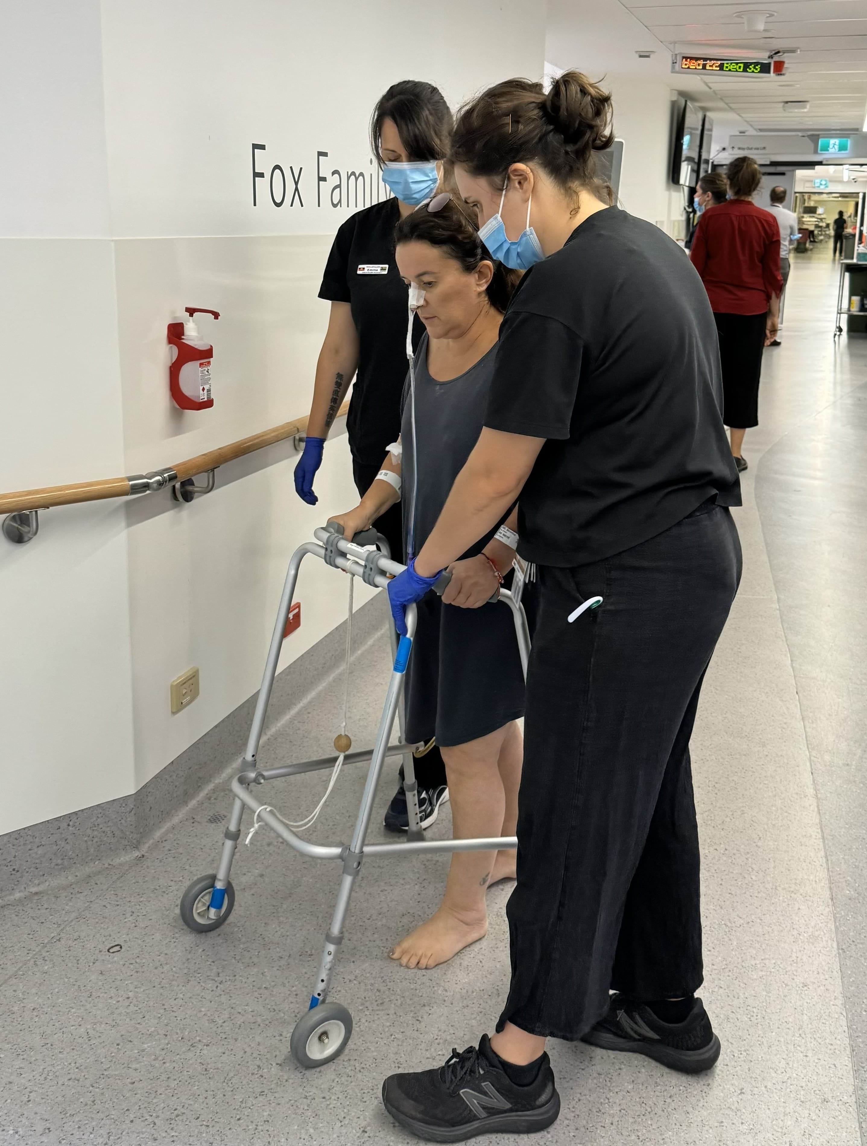 A woman using a walking frame and being assisted by two female nurses.