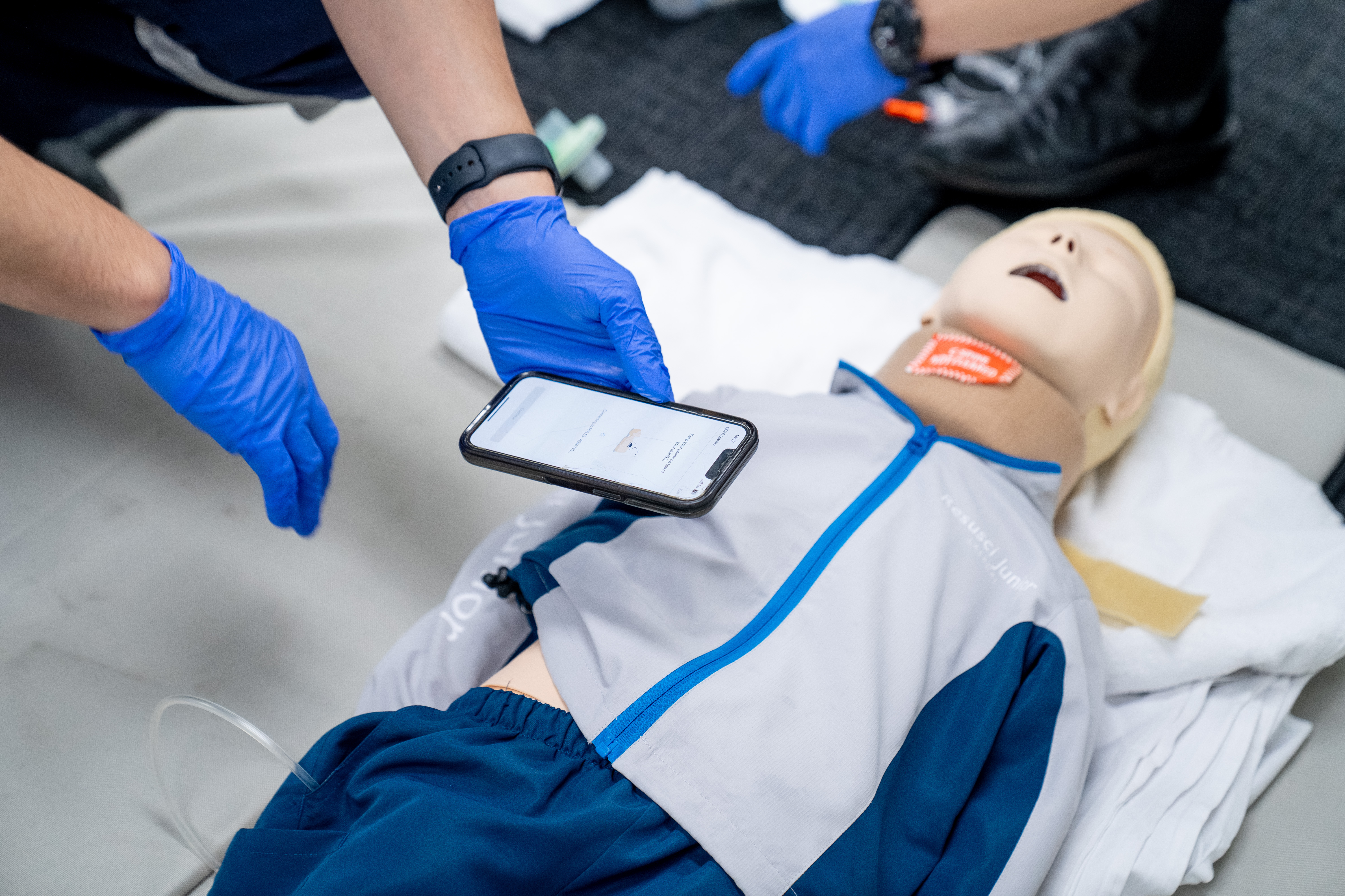 Someone's hand wearing a blue glove holding an iphone over the chest of a child sized manikin.
