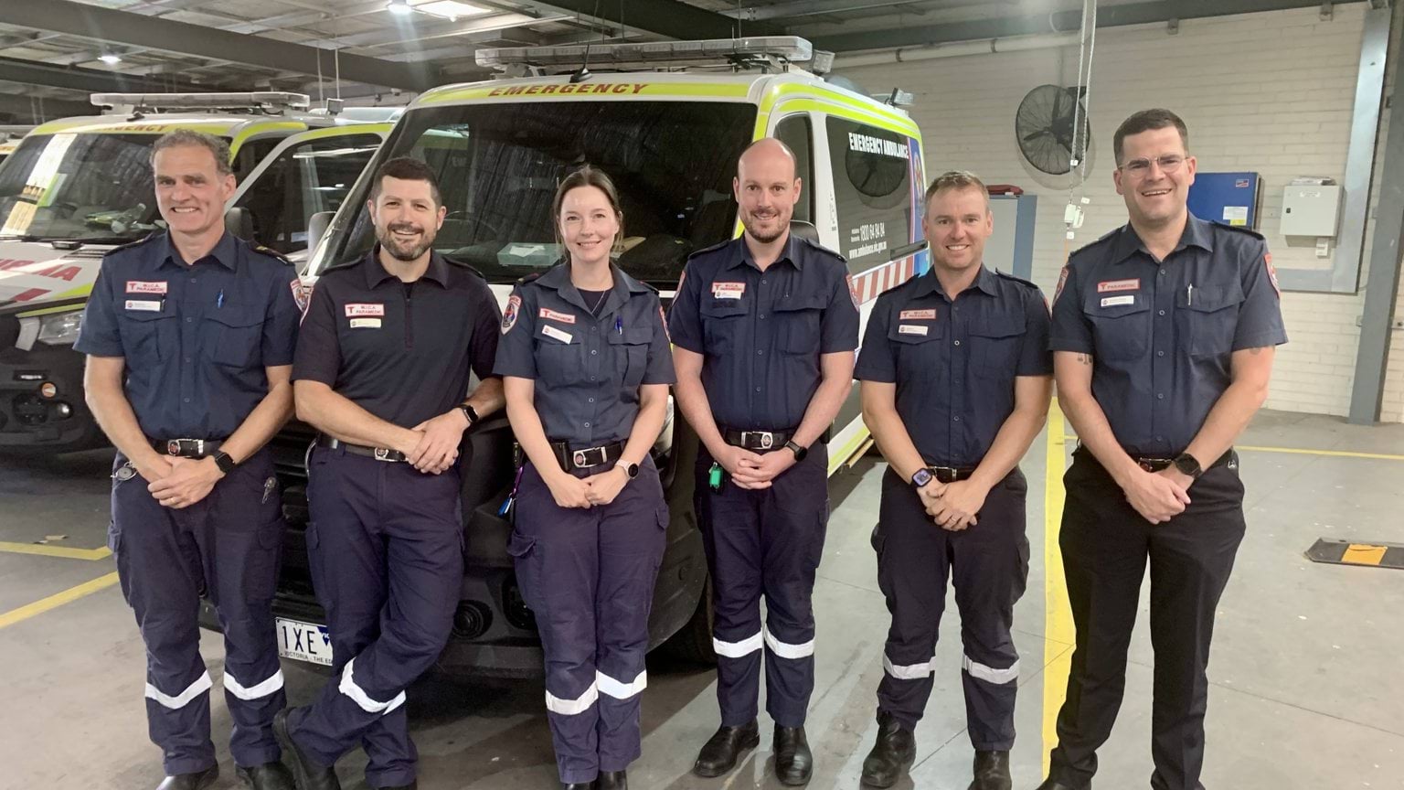 A group of six paramedics standing in uniform in front of an ambulance.