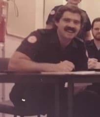 A male paramedic with a thick moustache sitting at a table.