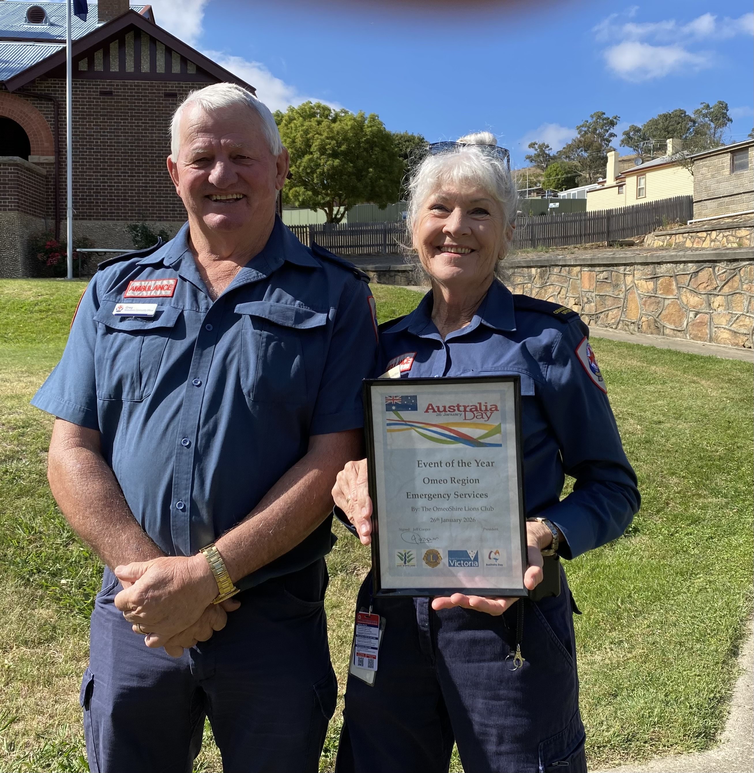 A man and a woman standing together in ambulance uniforms. The woman is holding a framed certificate. 