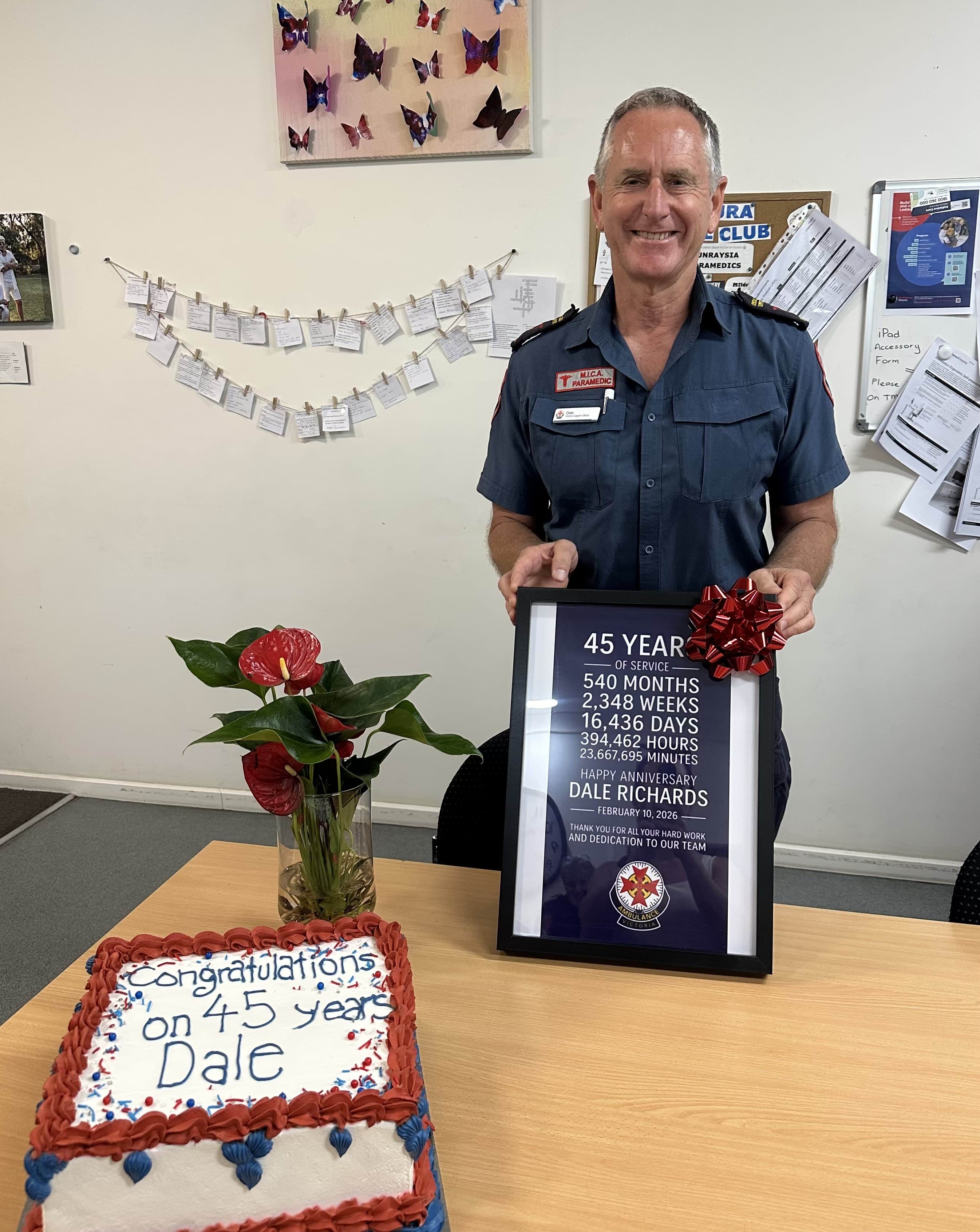 An Ambulance Victoria paramedic smiles holding a framed 45 year certificate. In front of him is a congratulatory cake.