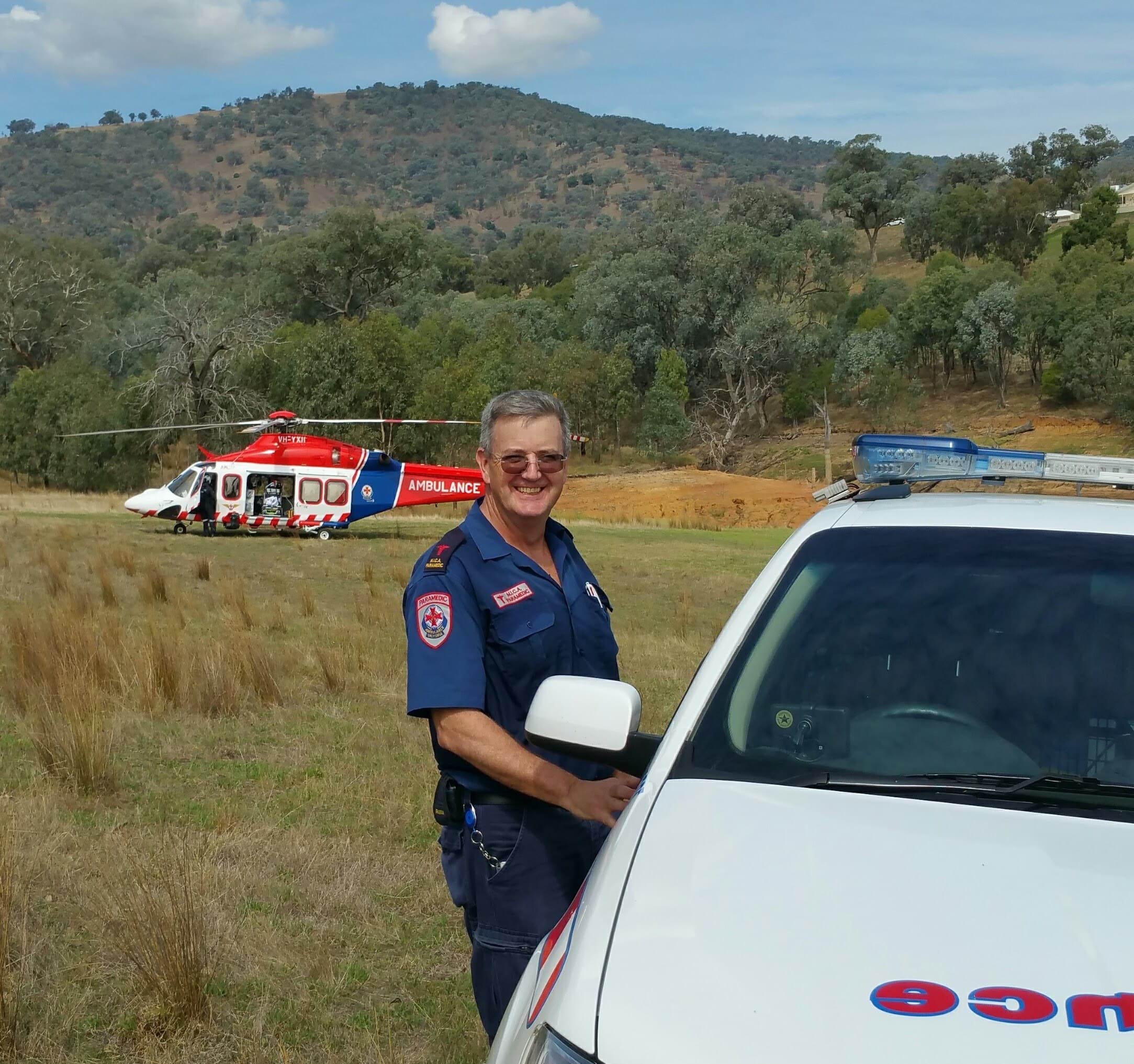 A man in a paramedic uniform standing next to a car. There is a helicopter in the background.