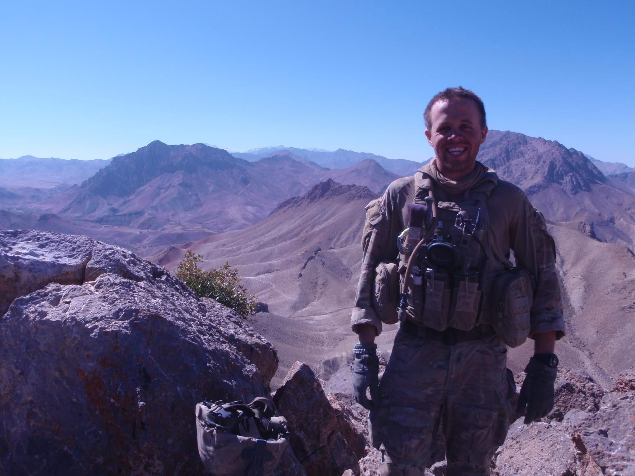 A soldier smiles for the camera in front of a landscape of mountains.
