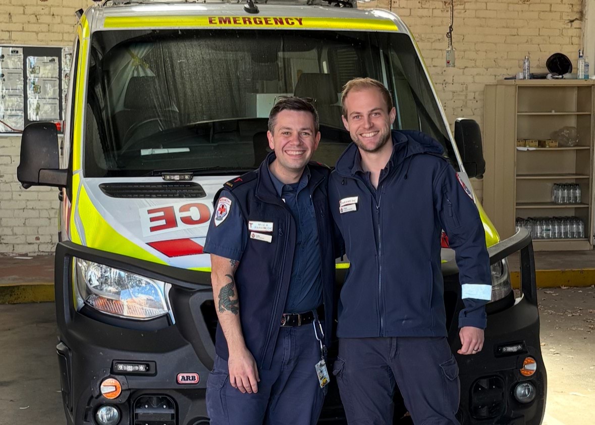Two paramedics smile in front of an ambulance.
