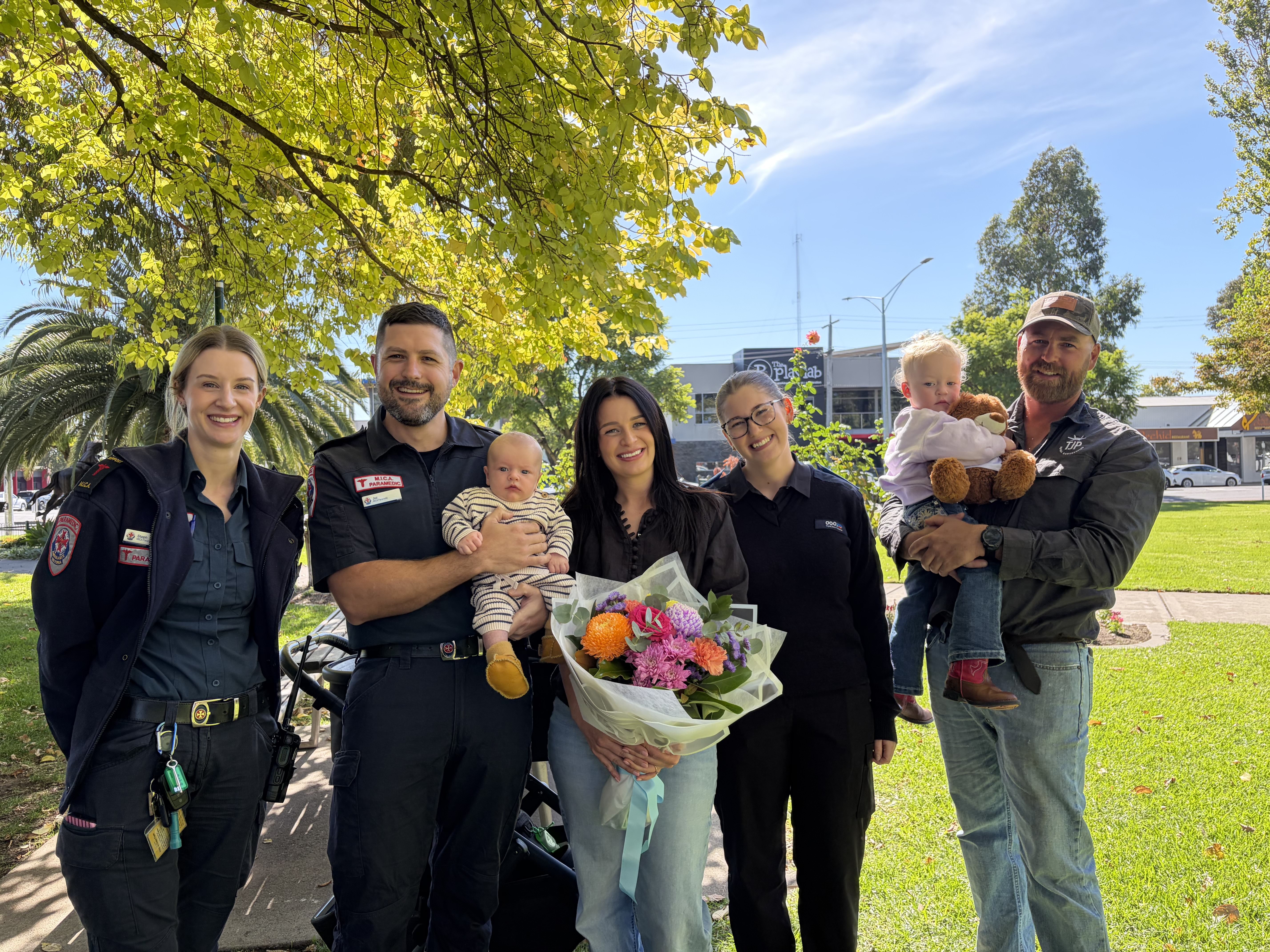 A group of people smile for the camera in a sunny park. A paramedic holds a baby.