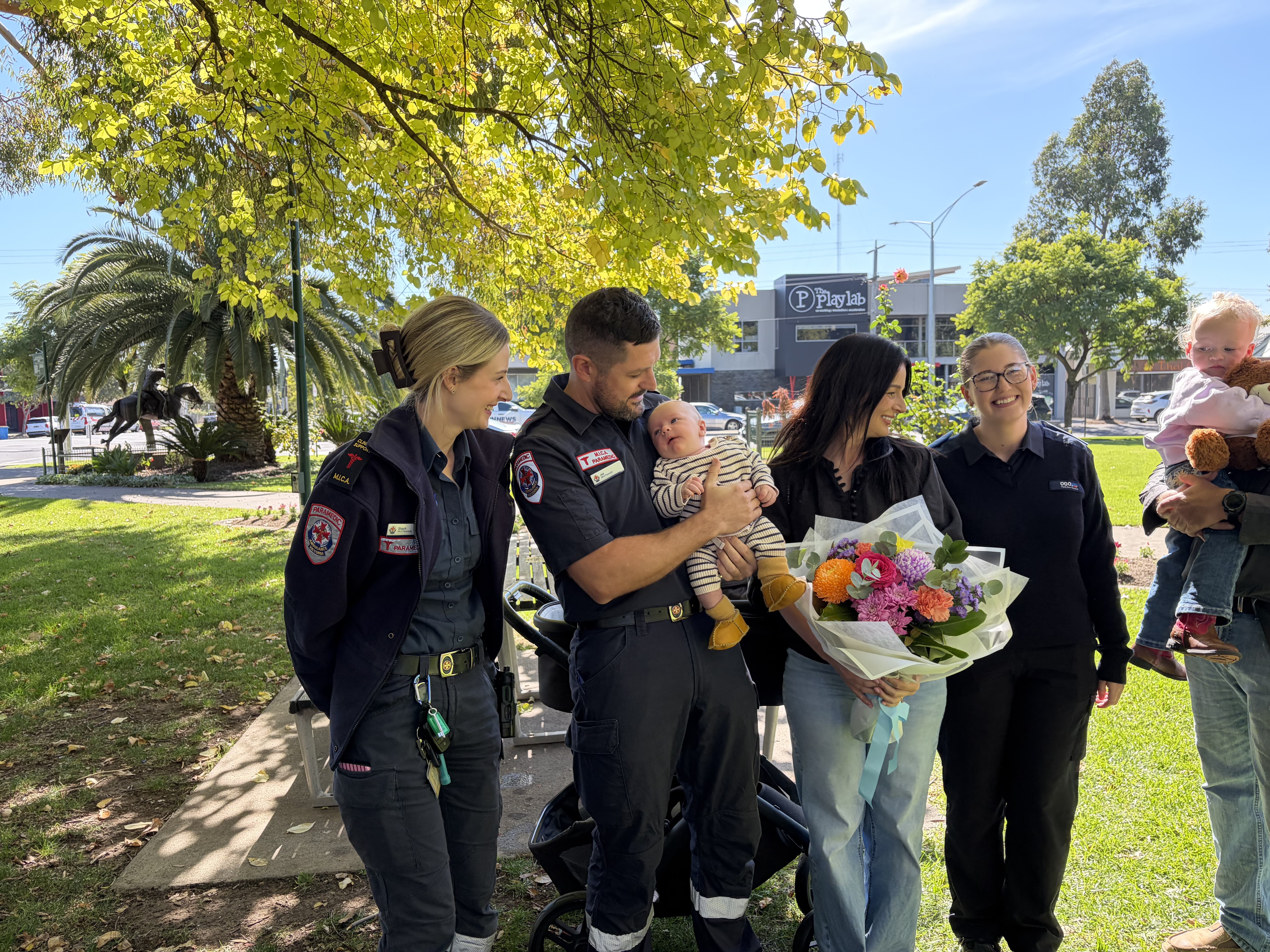 A group of people with a baby and a toddler in a sunny park.
