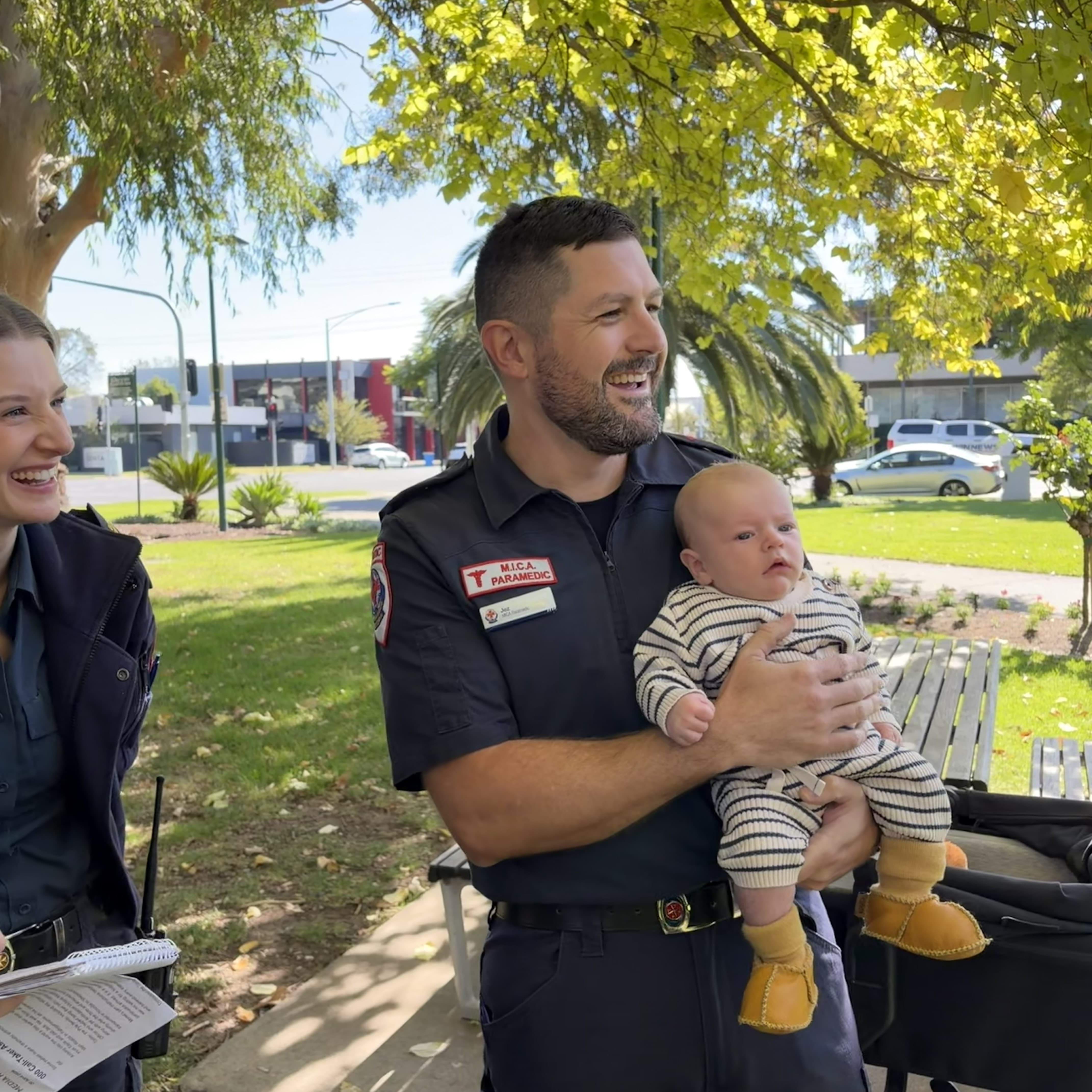 Paramedics smile at someone off camera. One of them is holding a baby.