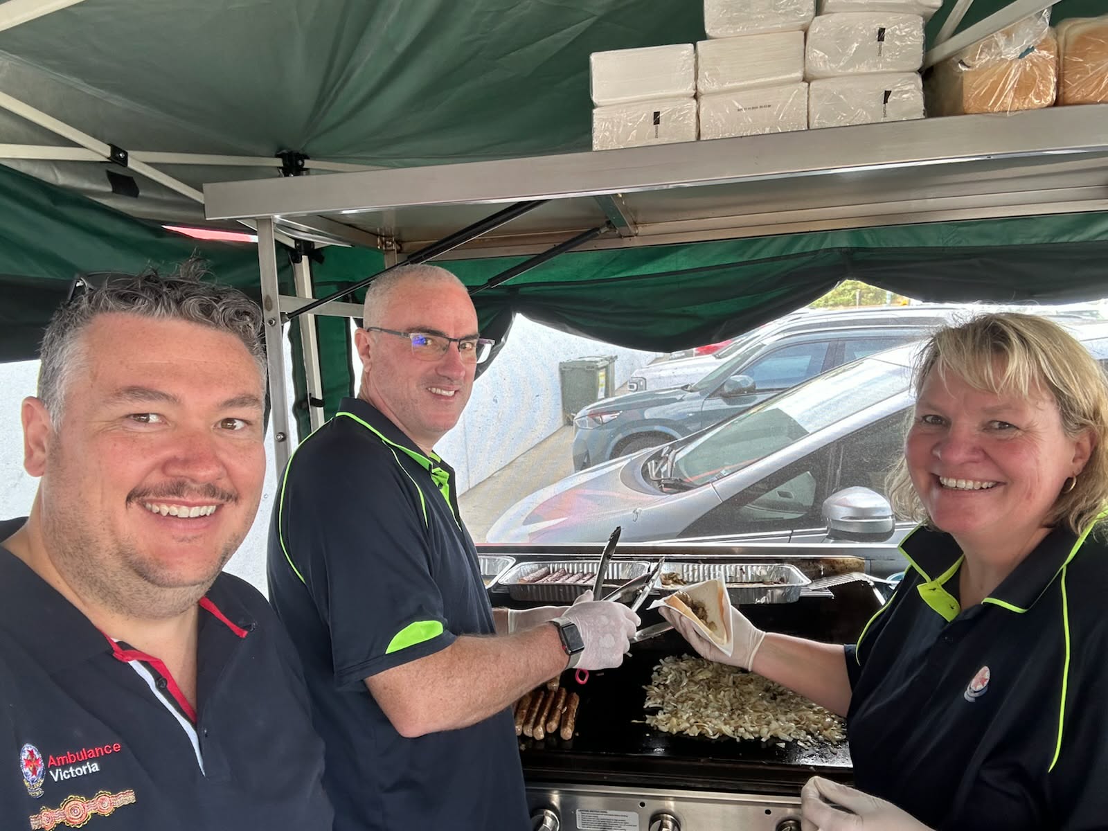 Three AV staff smile while cooking sausages and onions on a bbq.