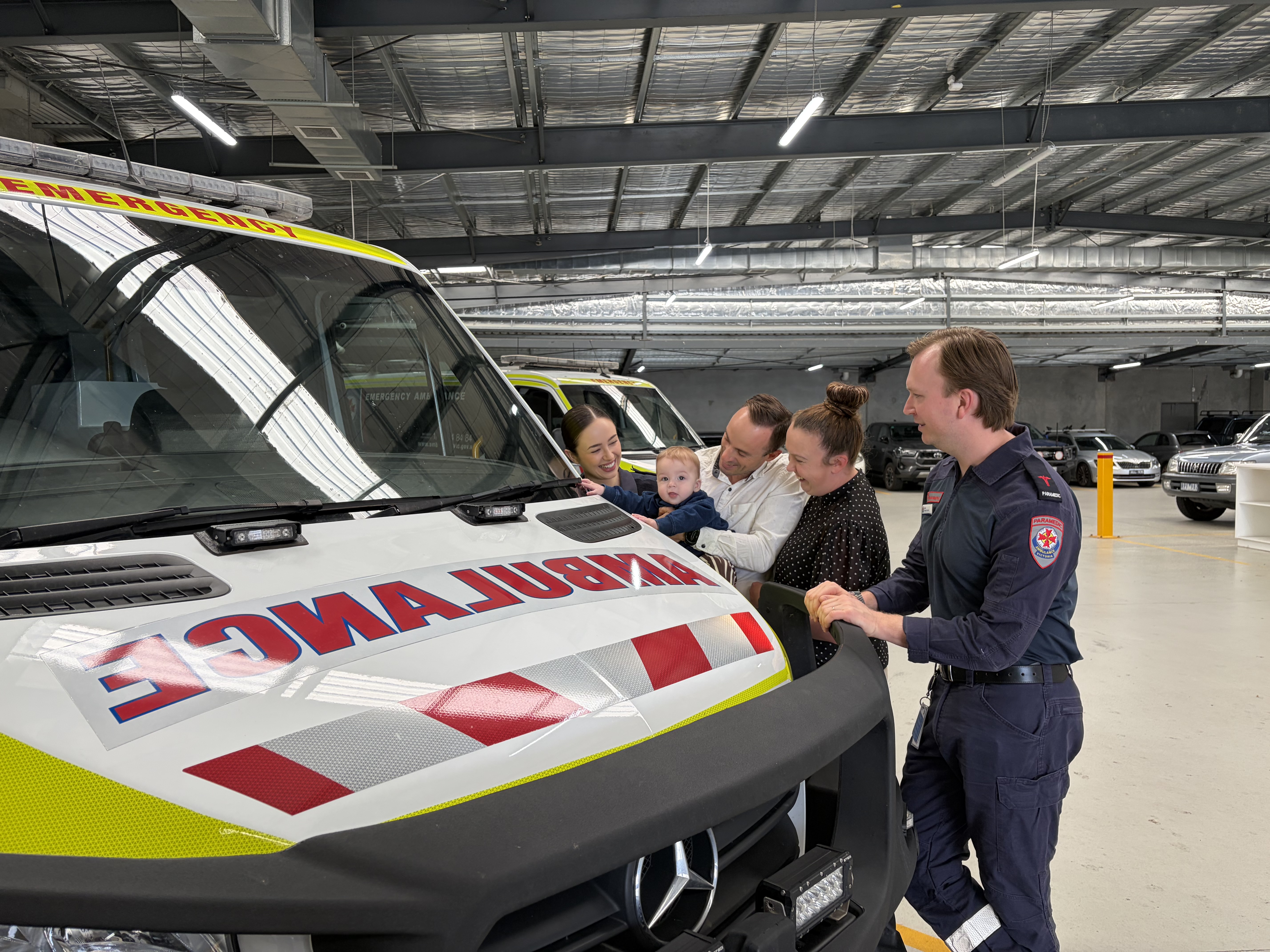 A baby puts his hands on the front of an ambulance. His parents and two paramedics smile at him.