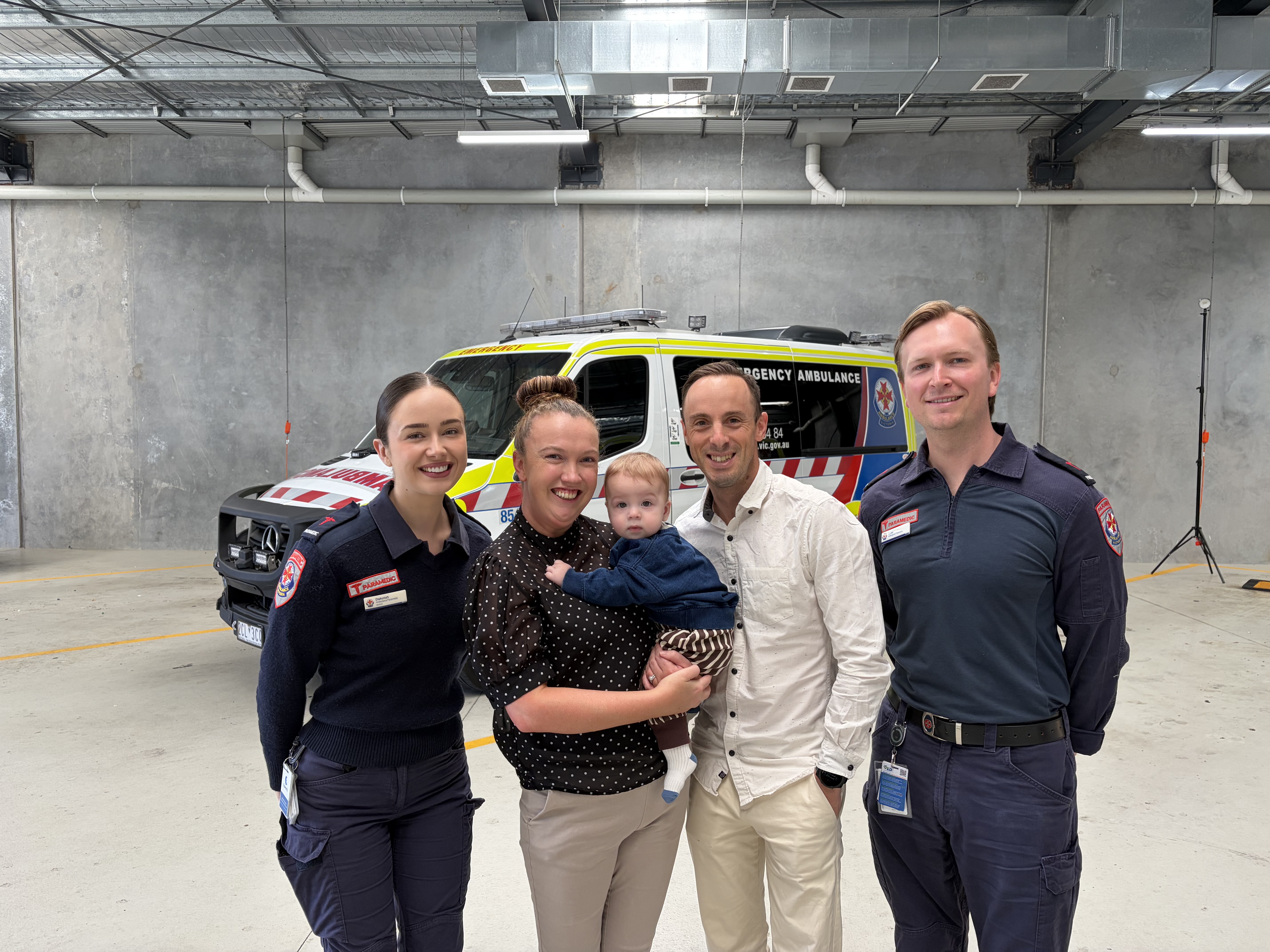 Two paramedics, two parents and a baby smile for the camera in front of an ambulance.