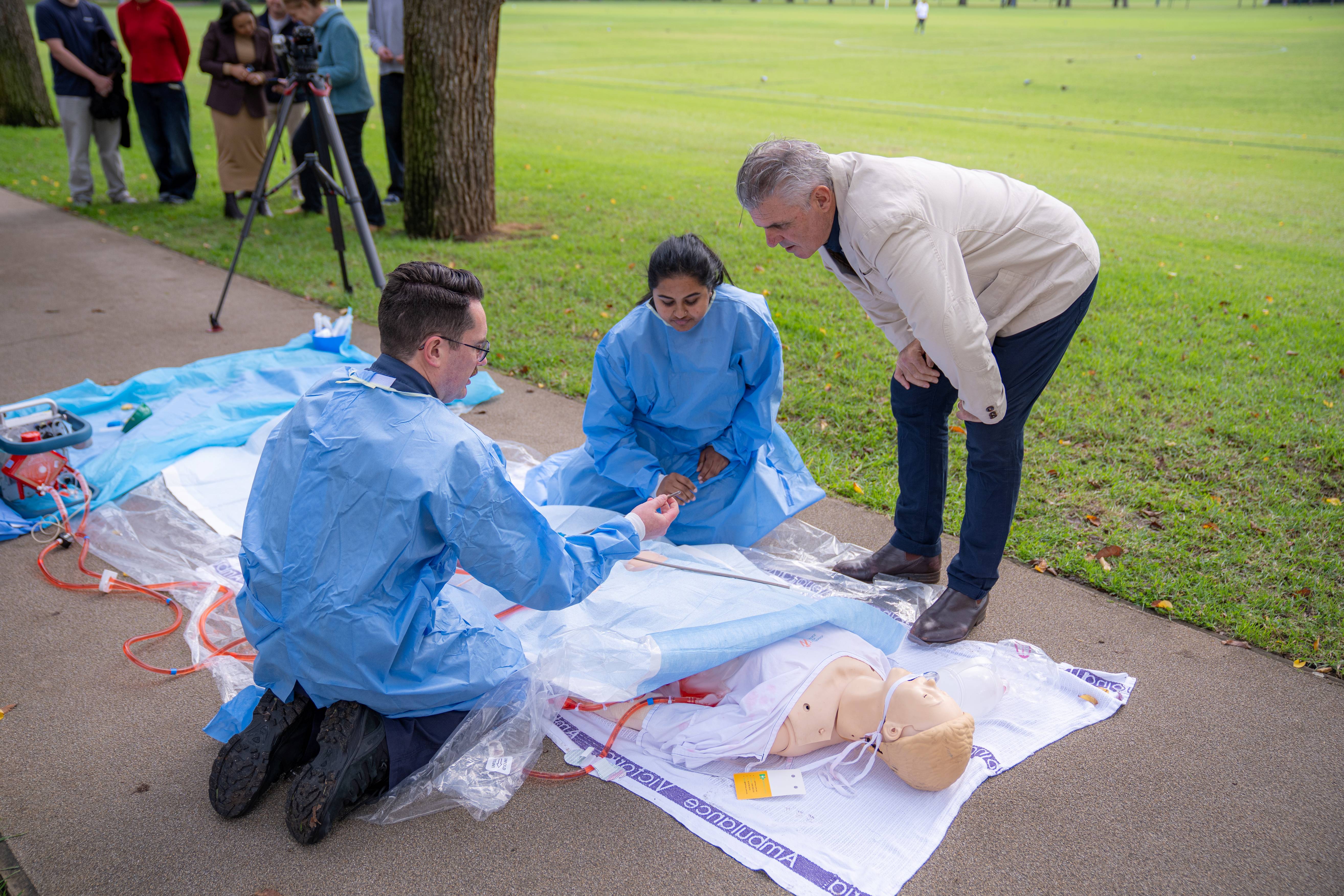 A medical demonstration on a path at a park, with a man watching on with interest.