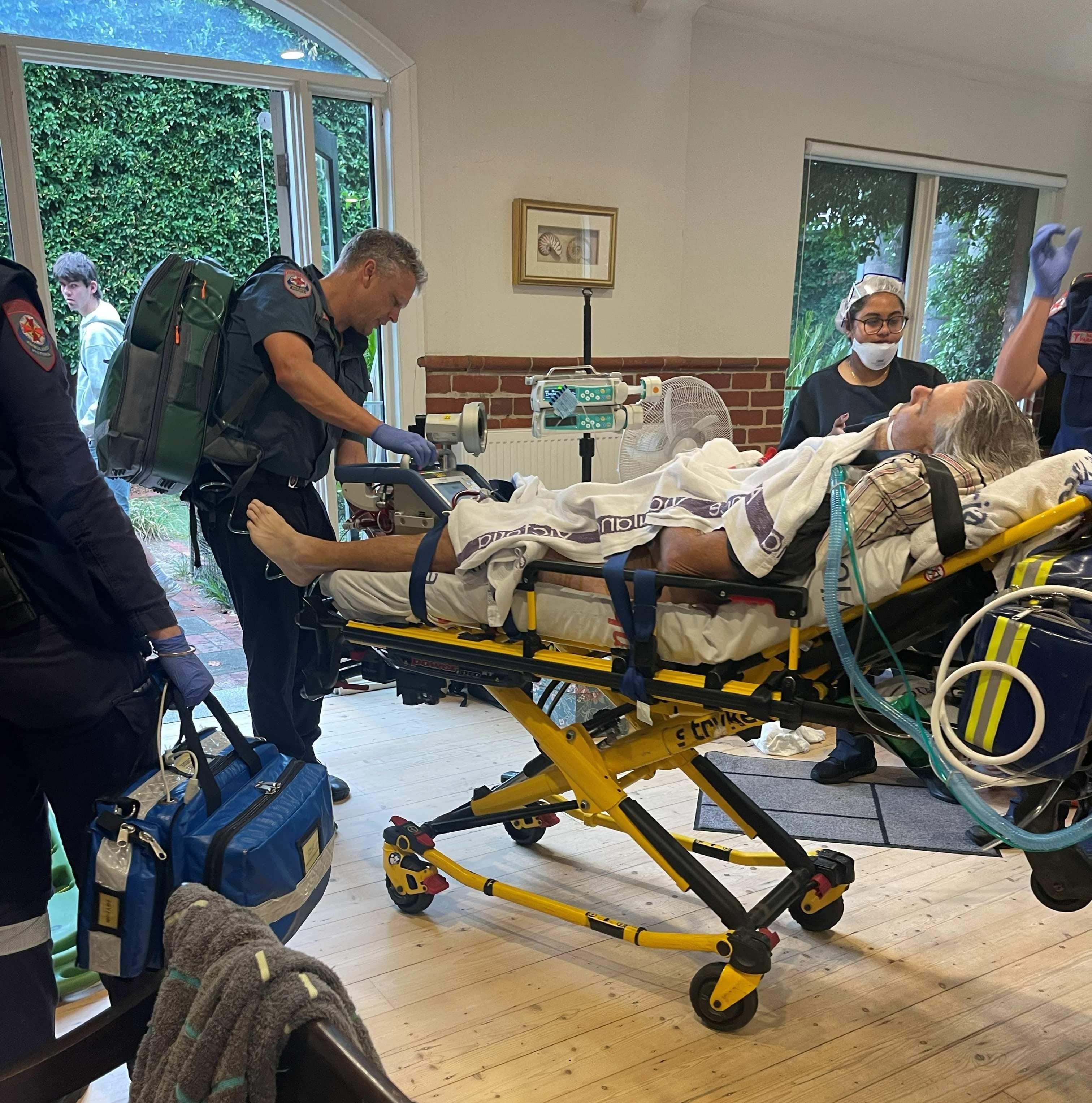 A man on an ambulance stretcher is wheeled out of his house by a paramedic.
