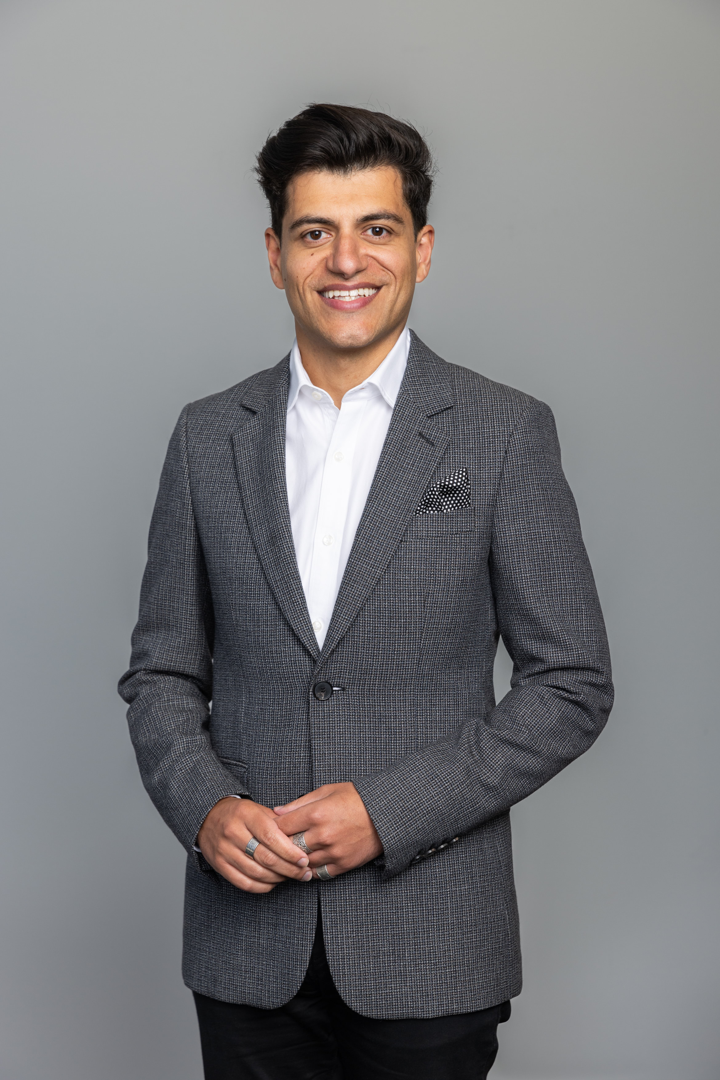 A man in corporate attire and a jacket smiling while his two hands are placed in the front.