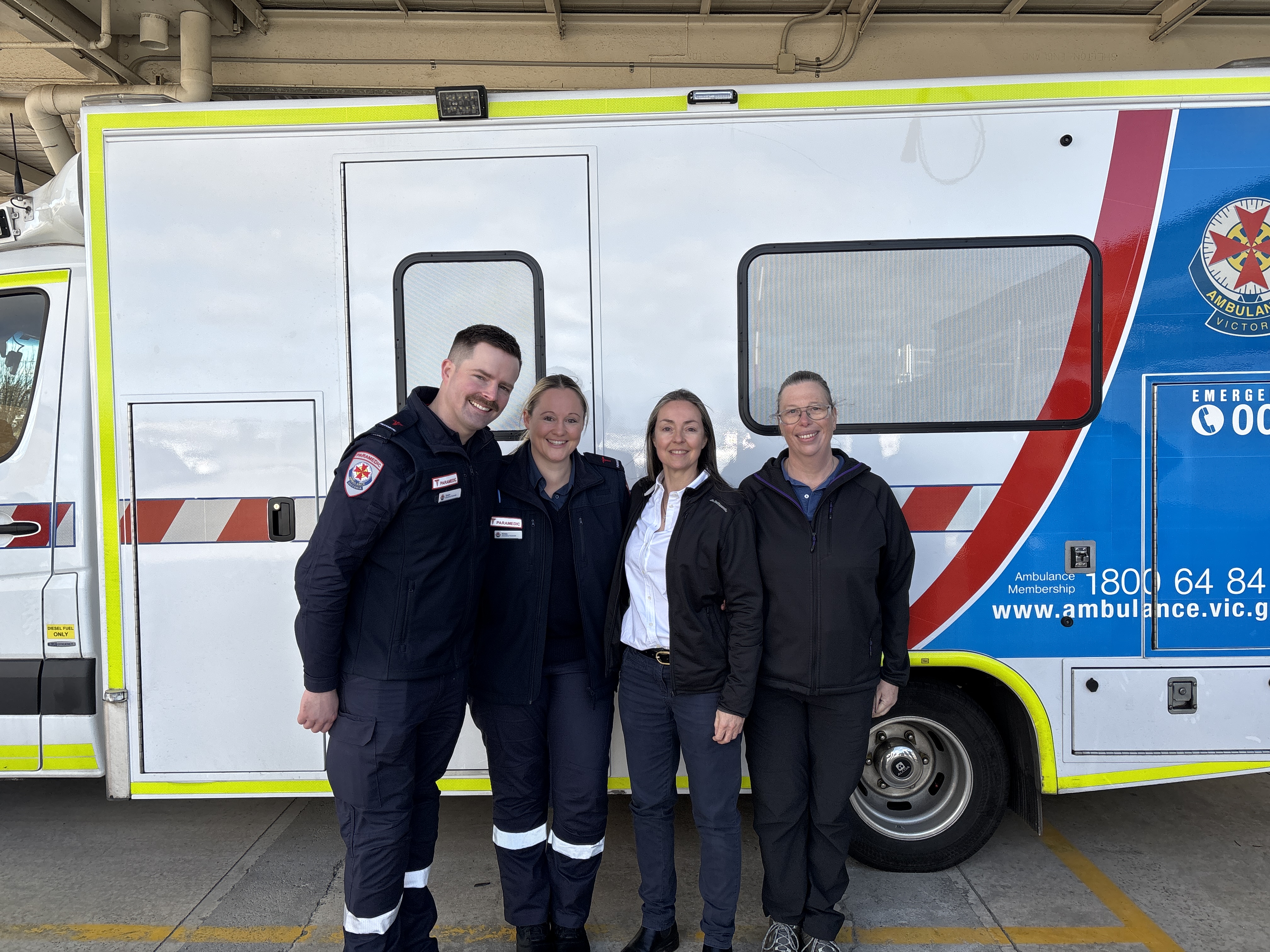 Two paramedics and two women standing in front of an ambulance while smiling at the camera.
