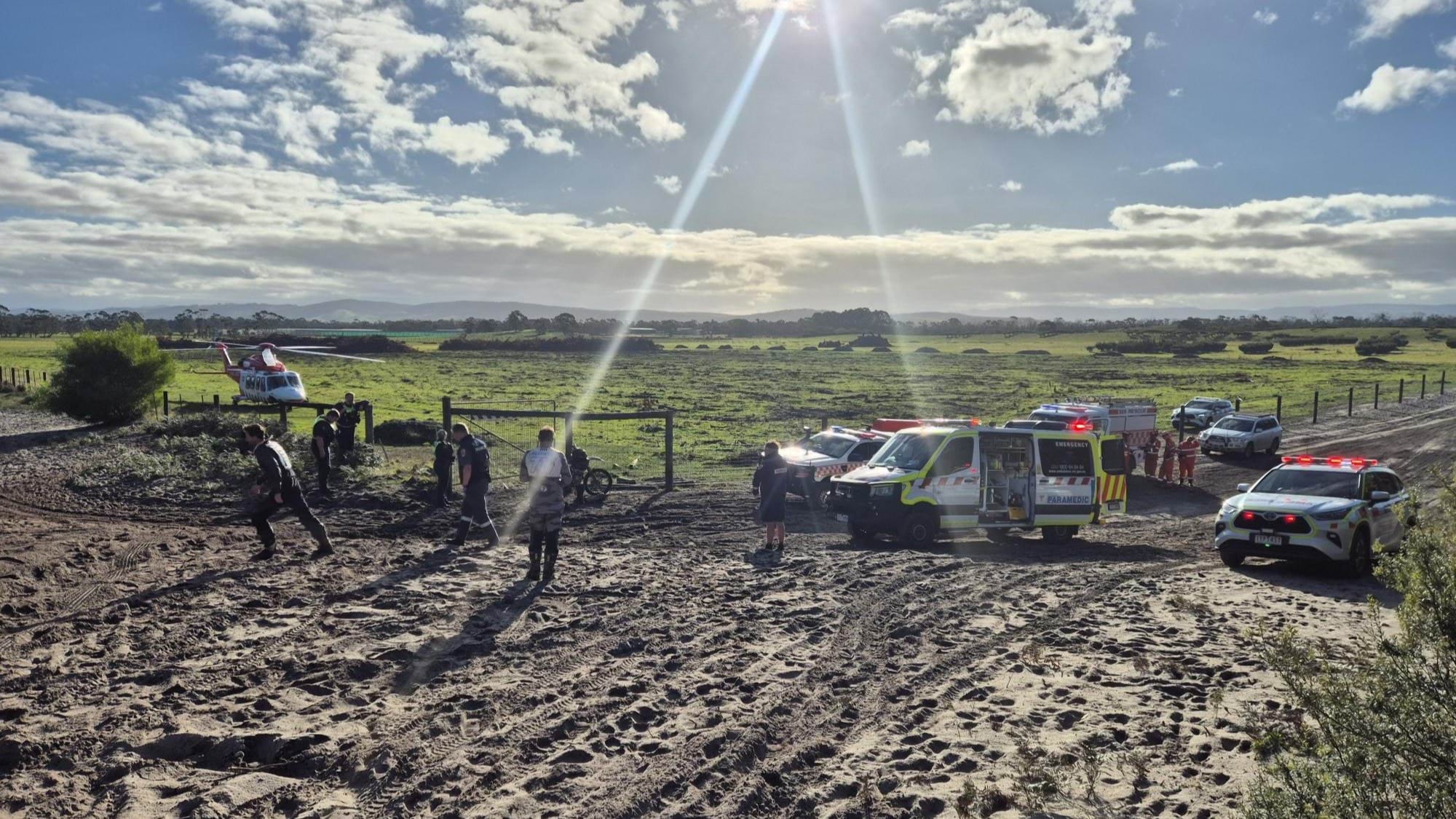 A paddock containing an ambulance helicopter with a dirt road to the right where two ambulance vehicles, a police car and SES vehicles are parked.