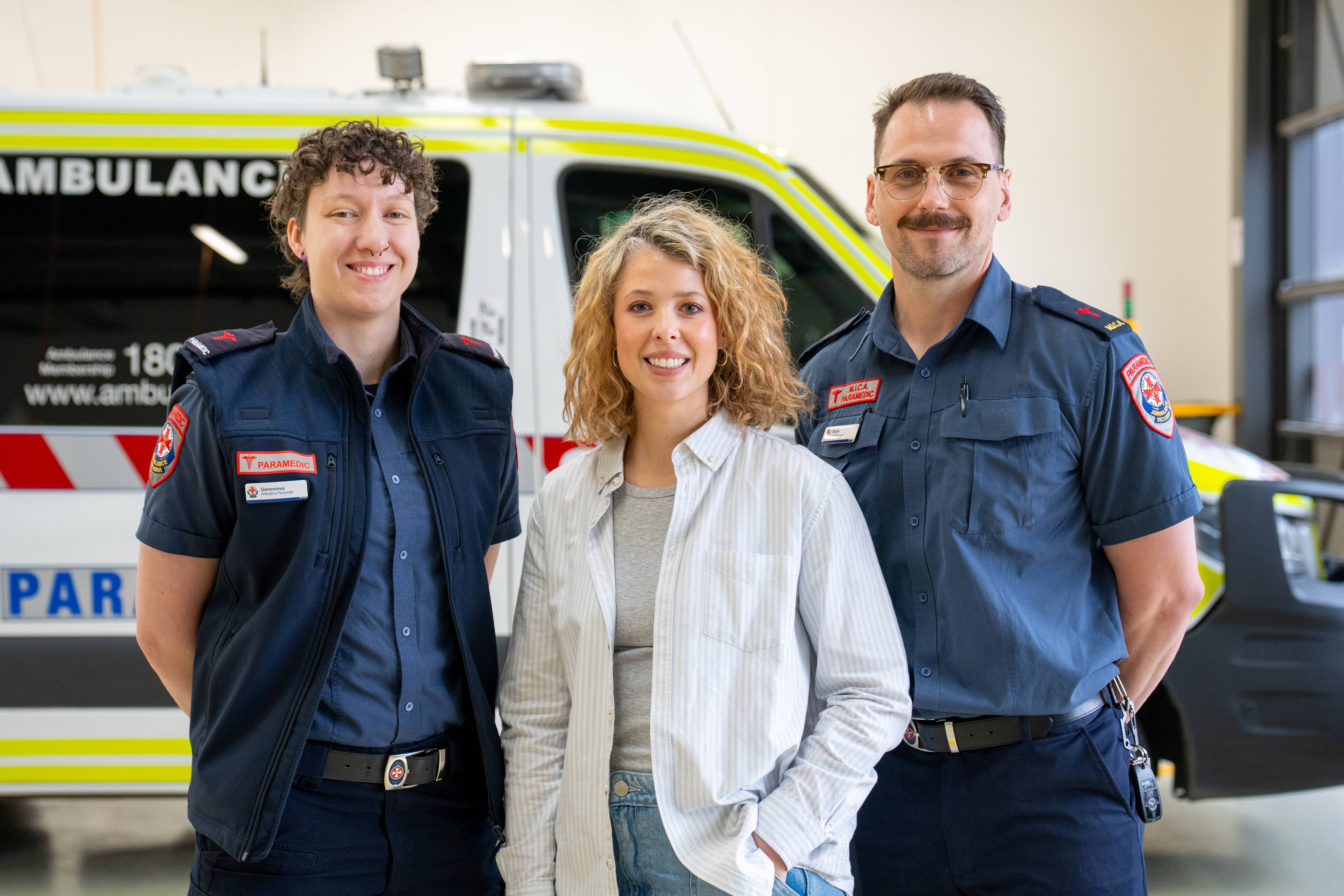 A woman smiles standing next to two paramedics.