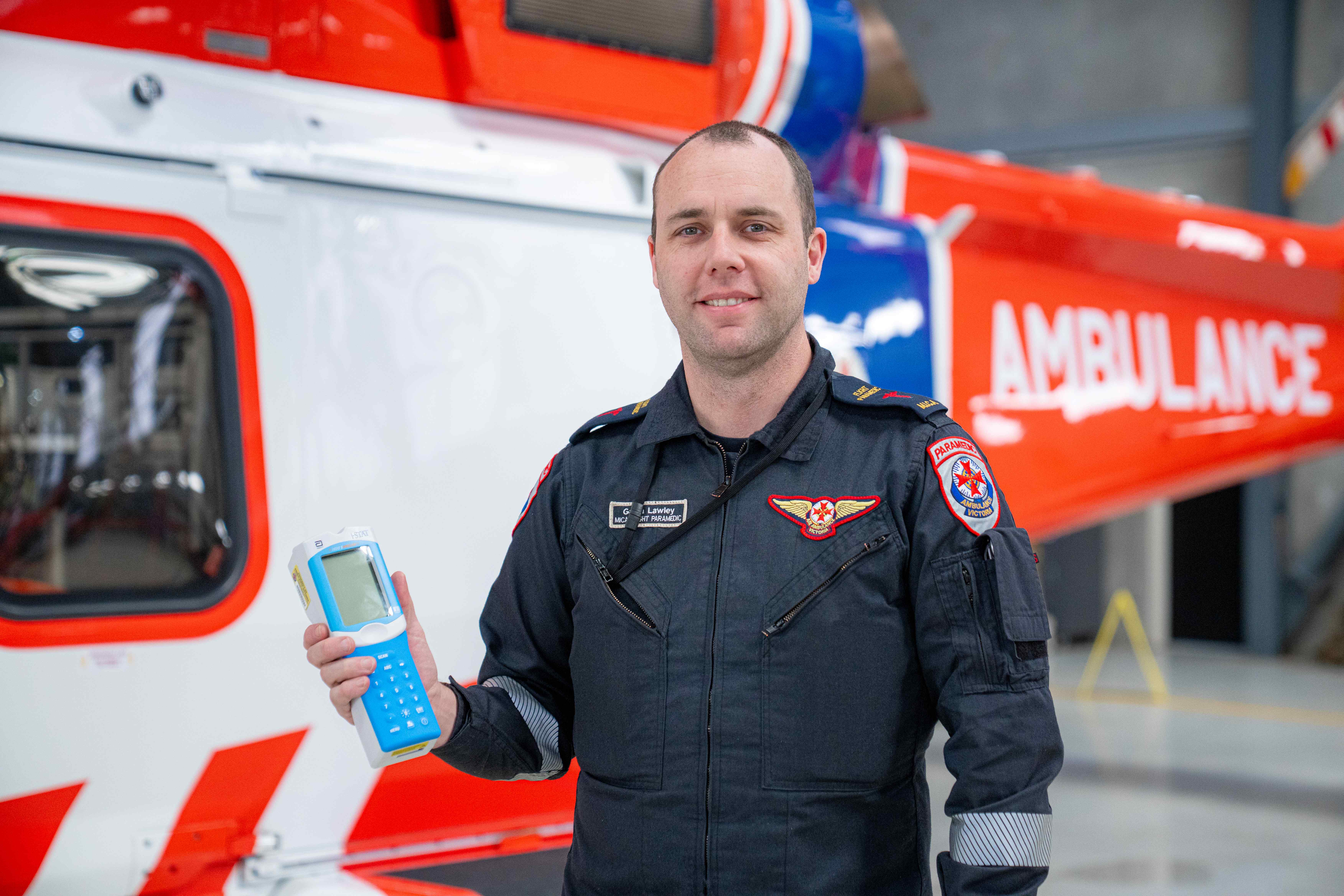 A paramedic holds a blue and grey handheld medical device. An air ambulance helicopter is behind him.