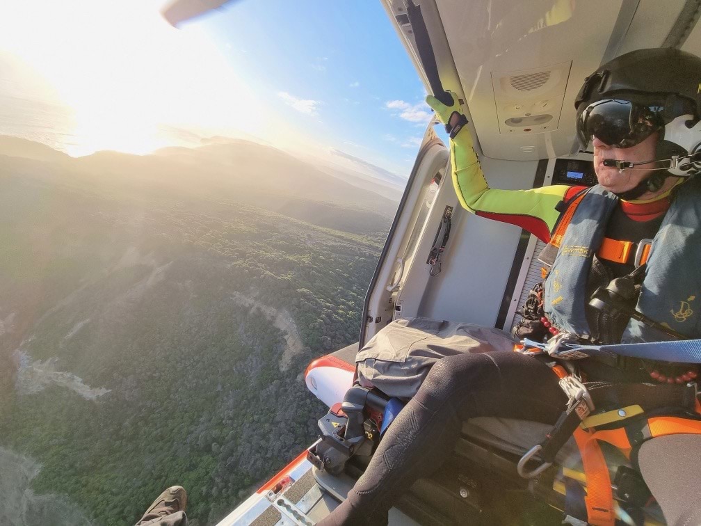 A MICA flight paramedic looks out the door of a helicopter flying over the coastline.