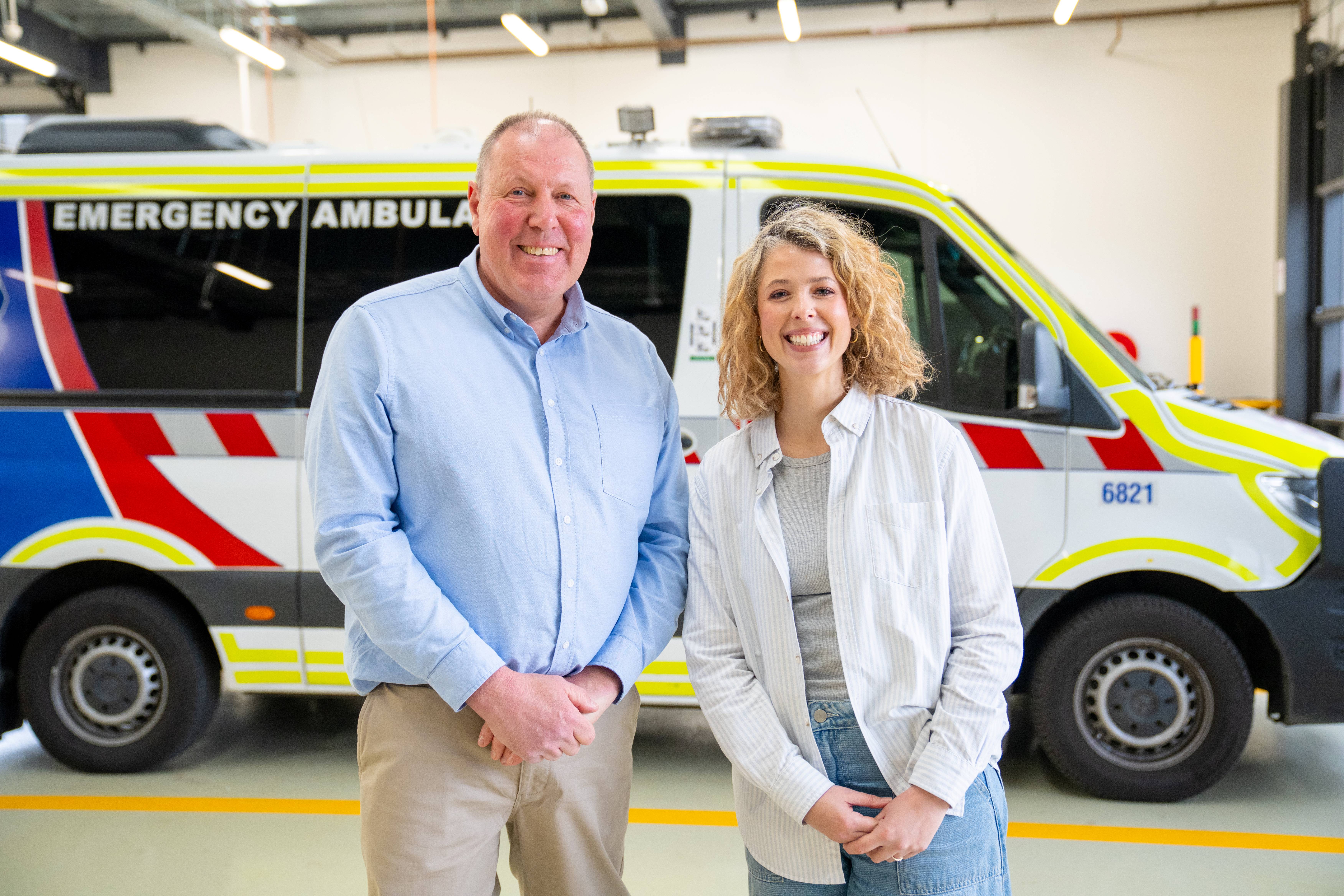 A man and woman smile for the camera in front of an ambulance.