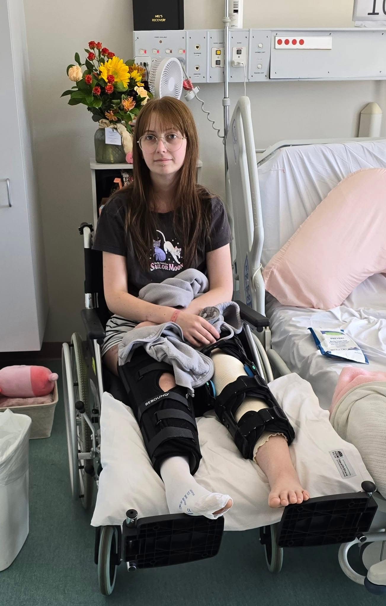 A patient with two leg braces sits in a wheelchair in a hospital room.