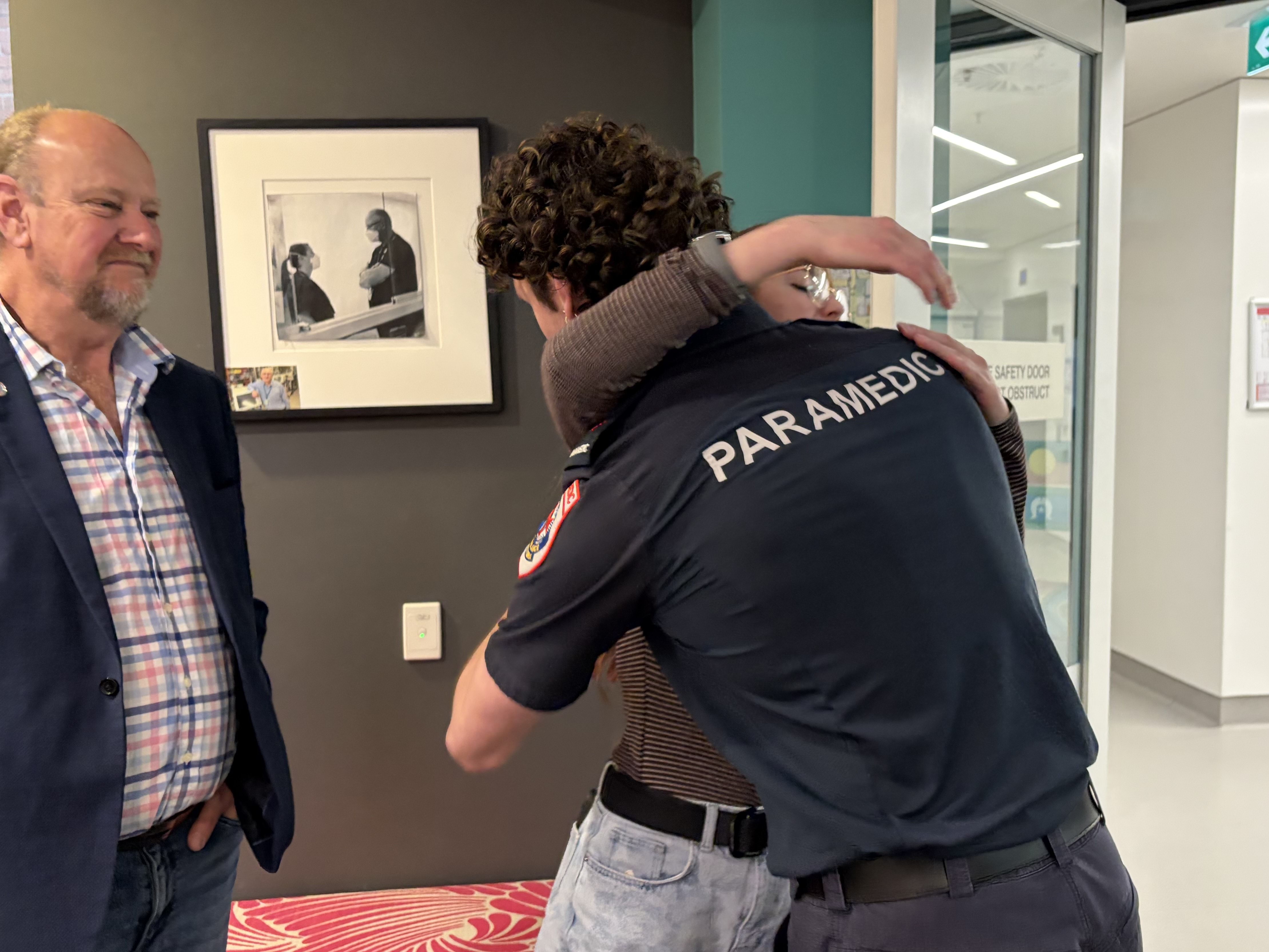 A paramedic hugs a woman. A man smiles.