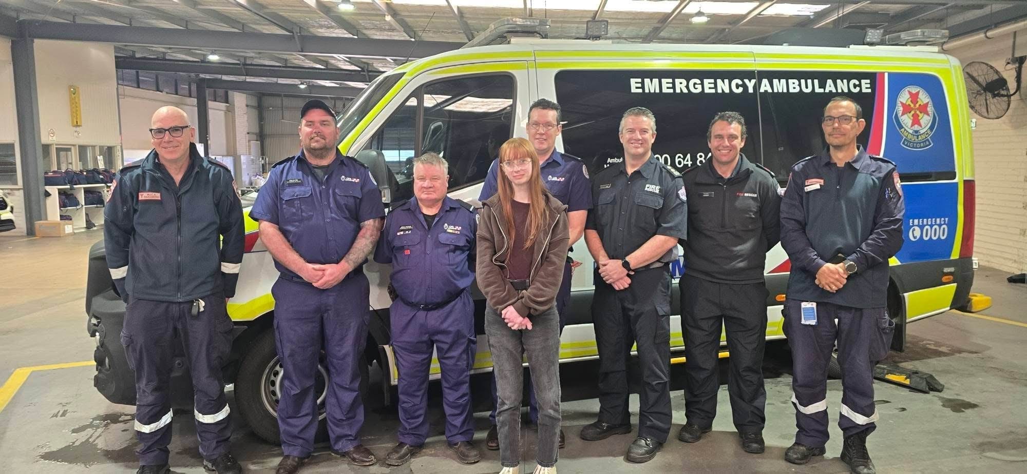 A young woman with a group of paramedics and firefighters in front of an ambulance.