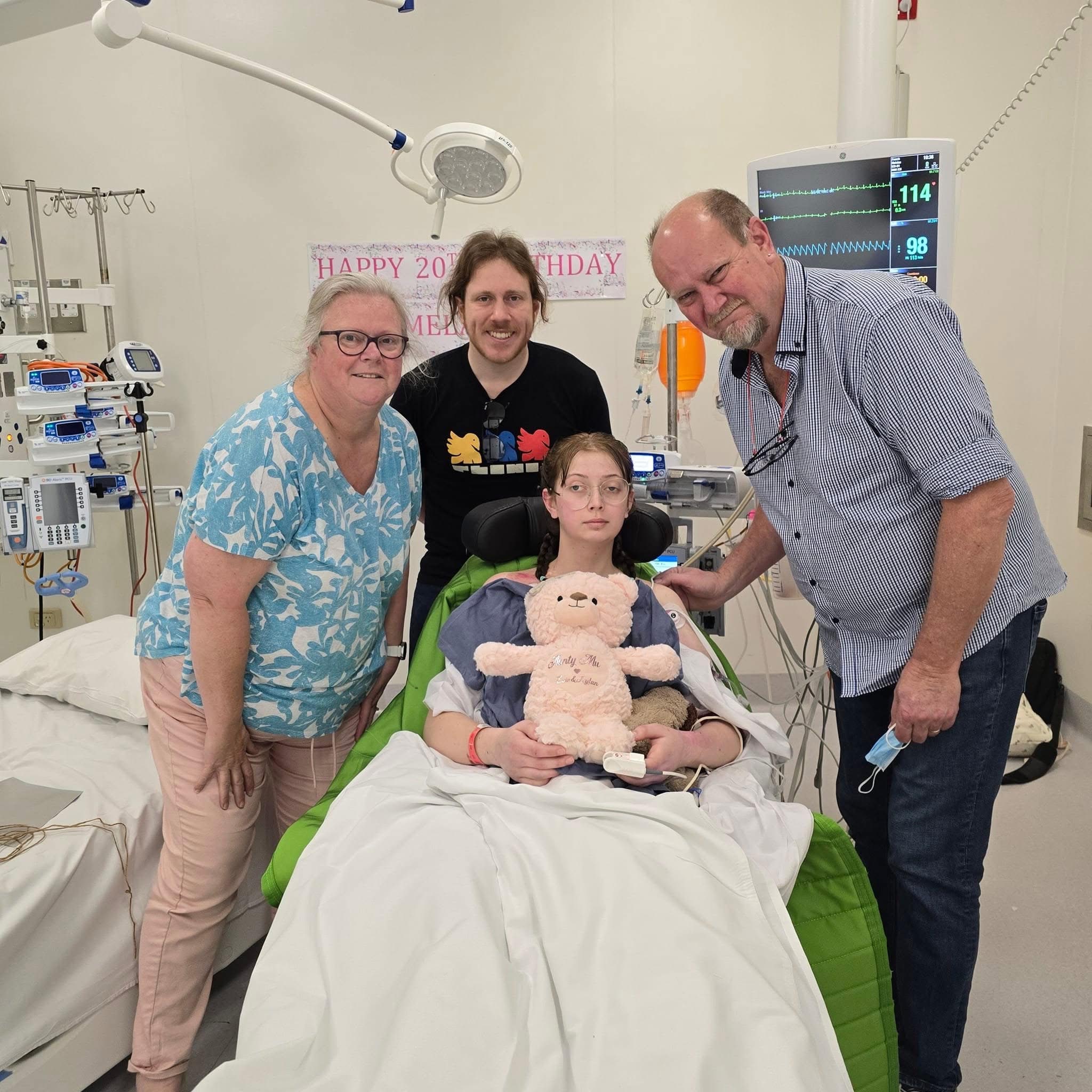 A patient in a hospital bed with three visitors. A 'happy birthday' sign is on the wall.