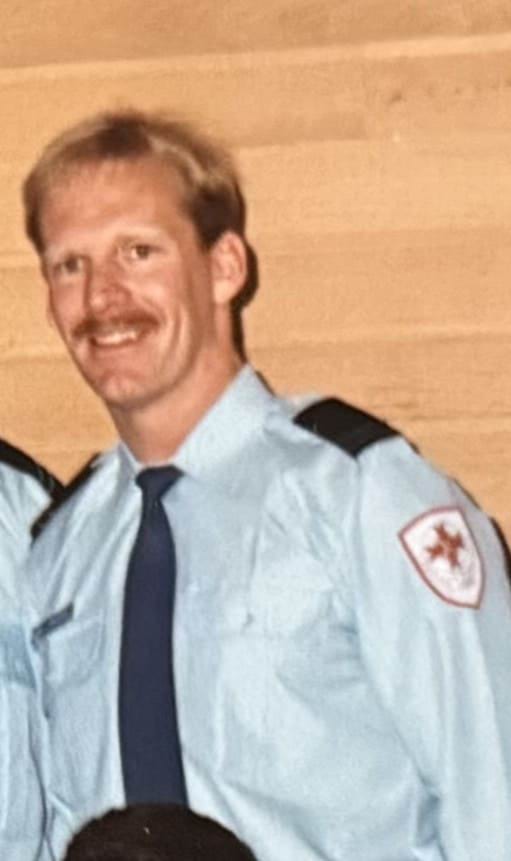 A man with blonde hair and a moustache wearing an ambulance shirt and tie.