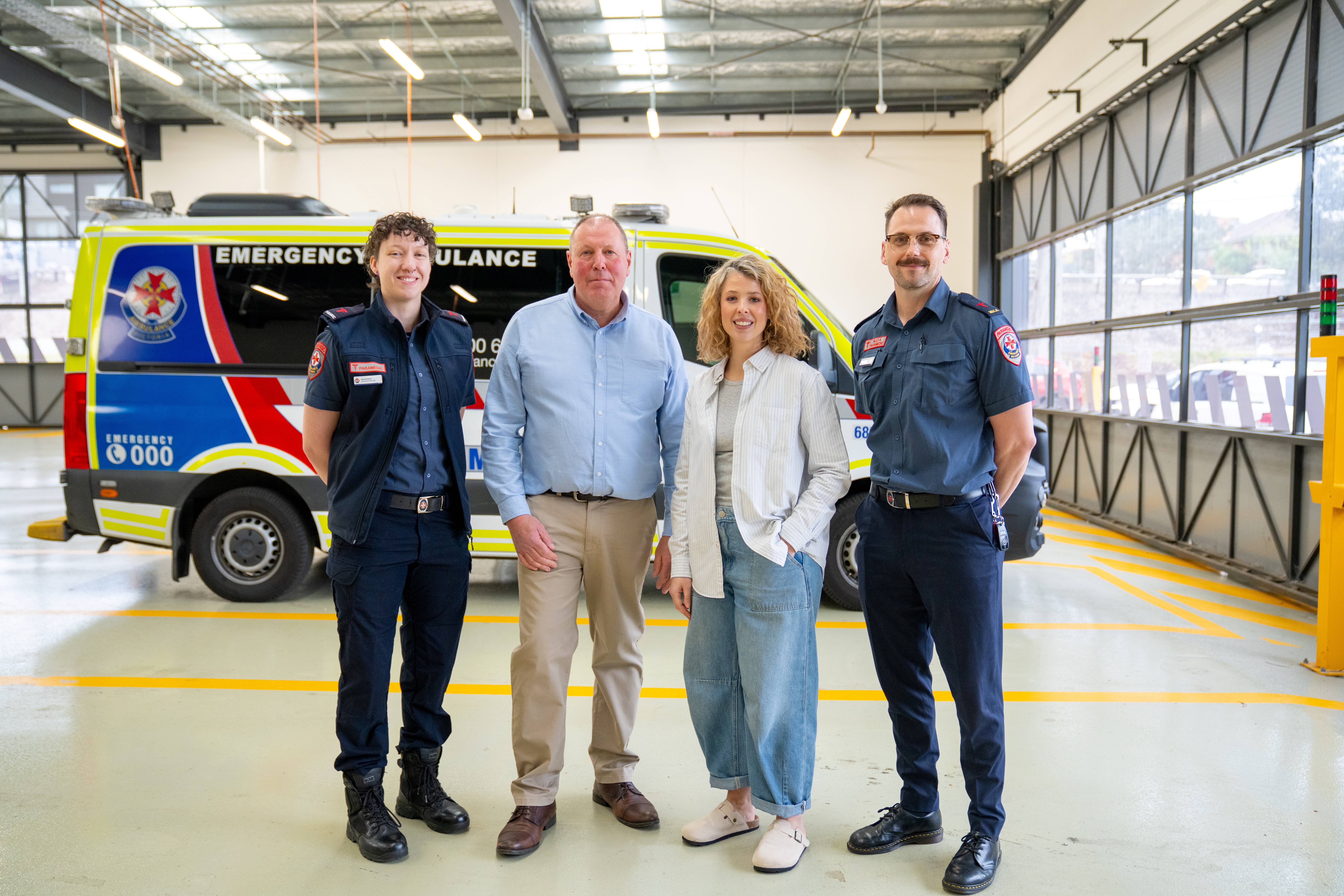 Two paramedics and a man and a woman smile in front of an ambulance.