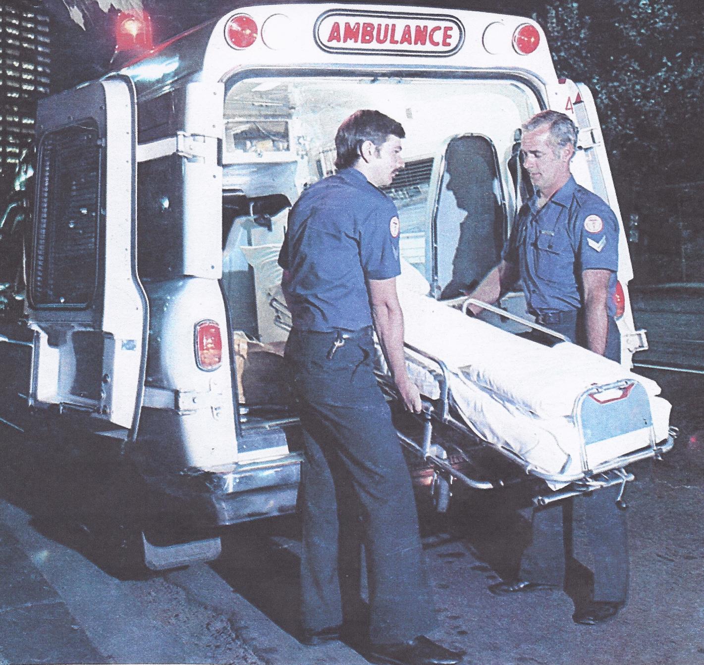 Two male paramedics taking a stretcher out of an ambulance. The photo is older so the ambulance is an older vehicle.