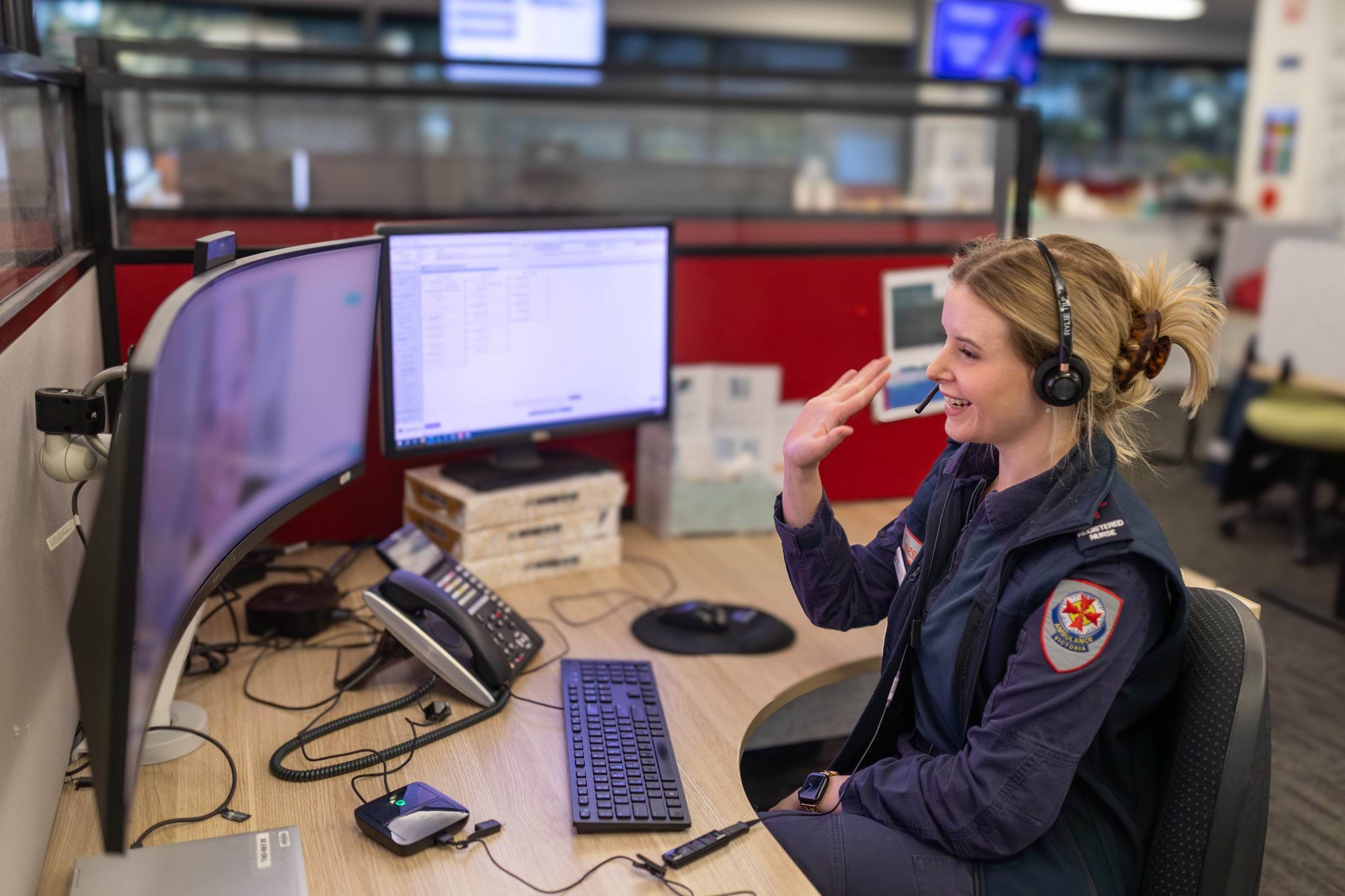 A person wearing a headset and a uniform with a patch featuring an AV red cross is seated at a work desk with multiple computer monitors. They are looking at a screen and gesturing with a wave of their hand. The setting is an office environment.