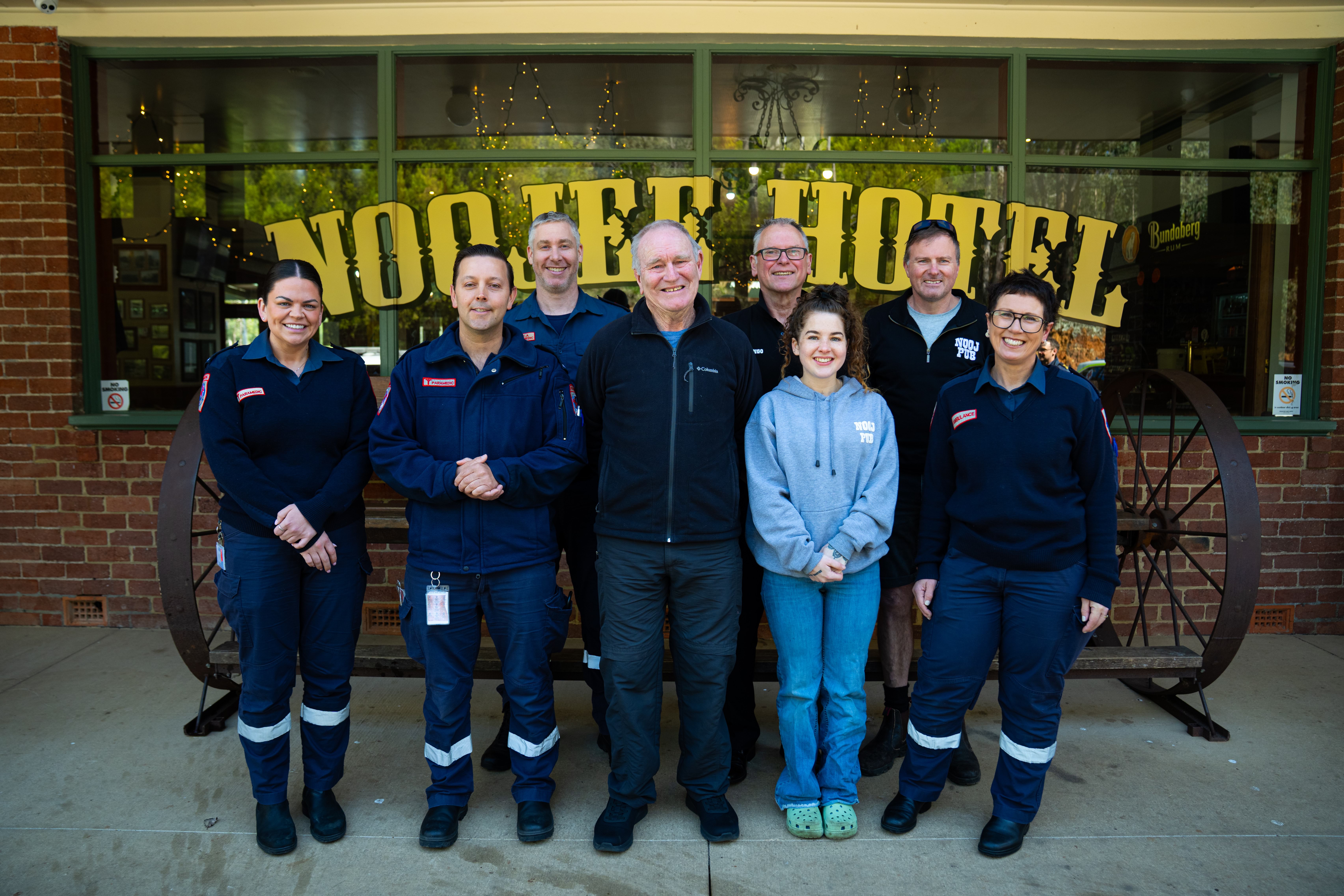 A group of eight smiling people standing in front of a sign on a window that says 'Noojee Hotel'.