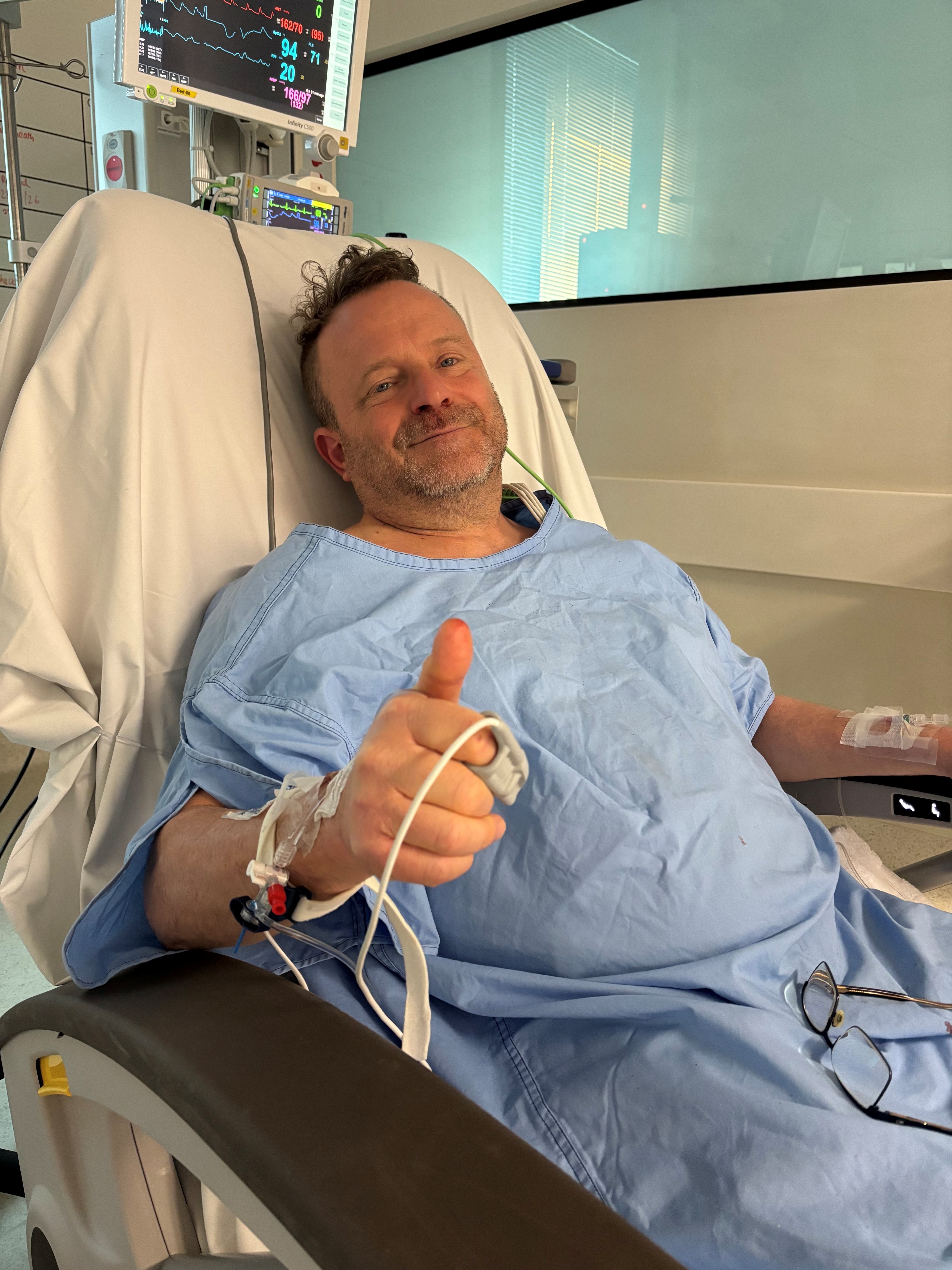 Adam in a hospital bed, dressed in a blue hospital gown giving a thumbs up gesture
