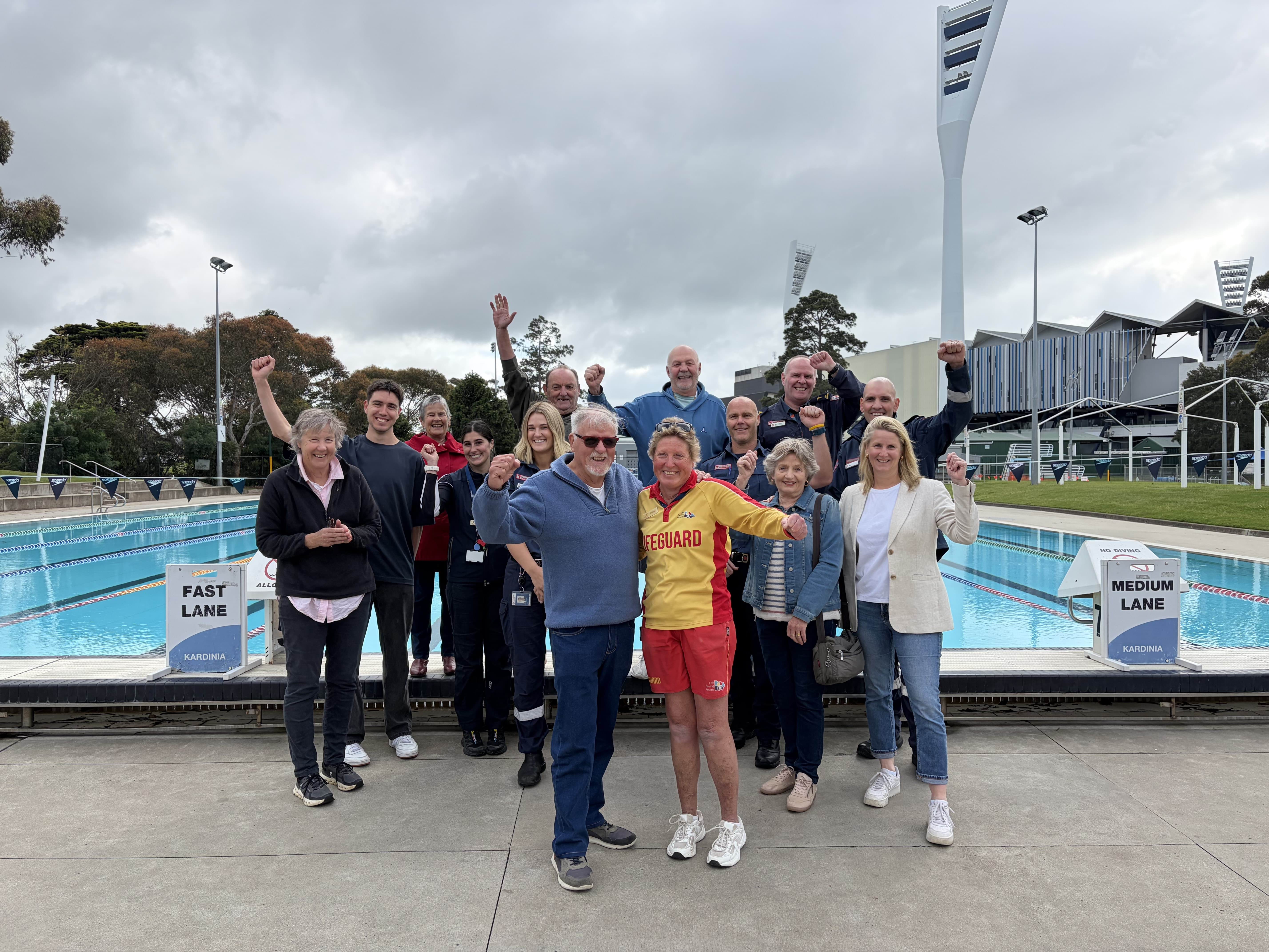 A group of people including paramedics and a lifeguard cheer in front of an outdoor swimming pool.
