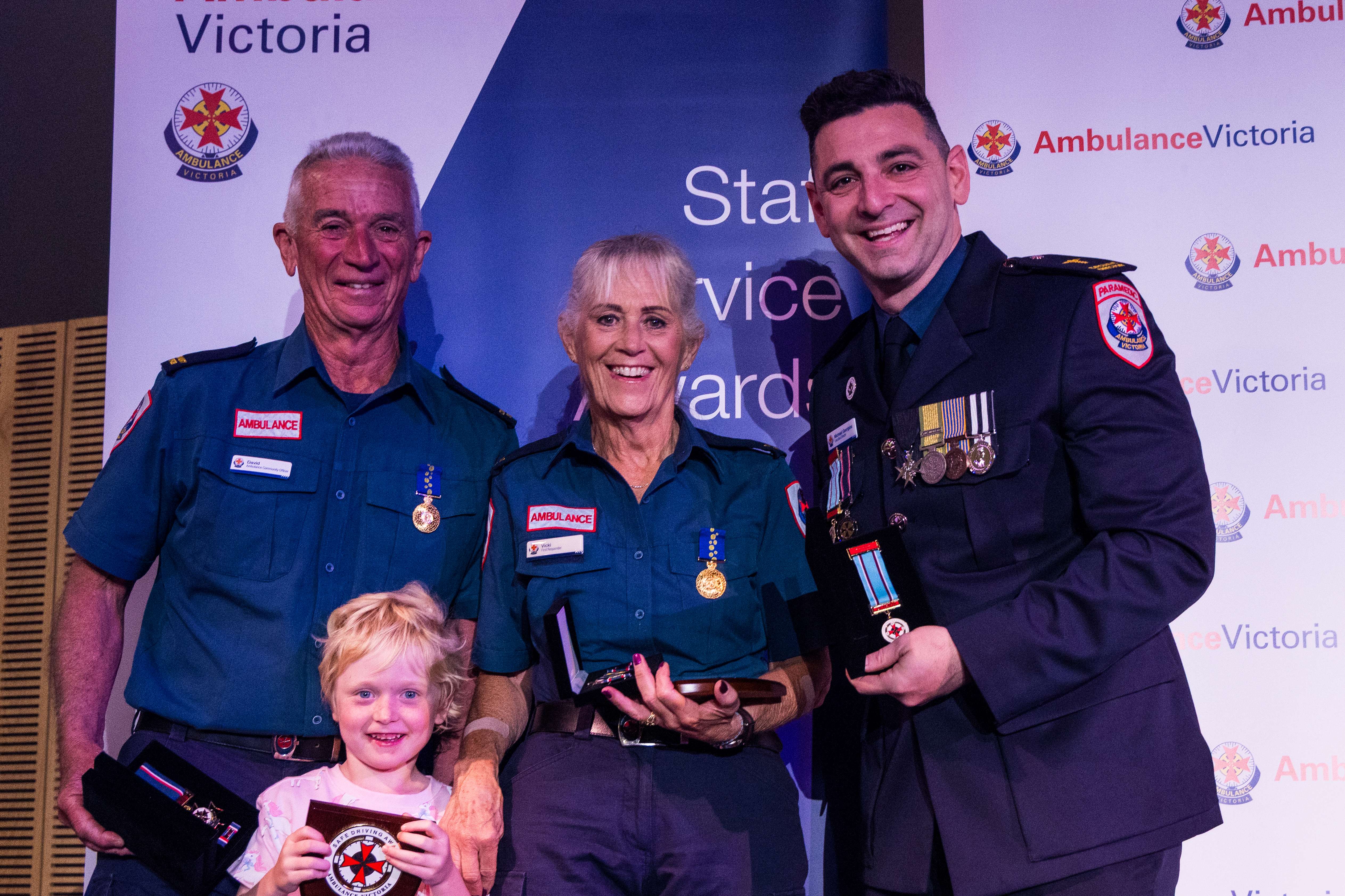 Three adults and a child smile at an Ambulance Victoria awards event, holding medals.