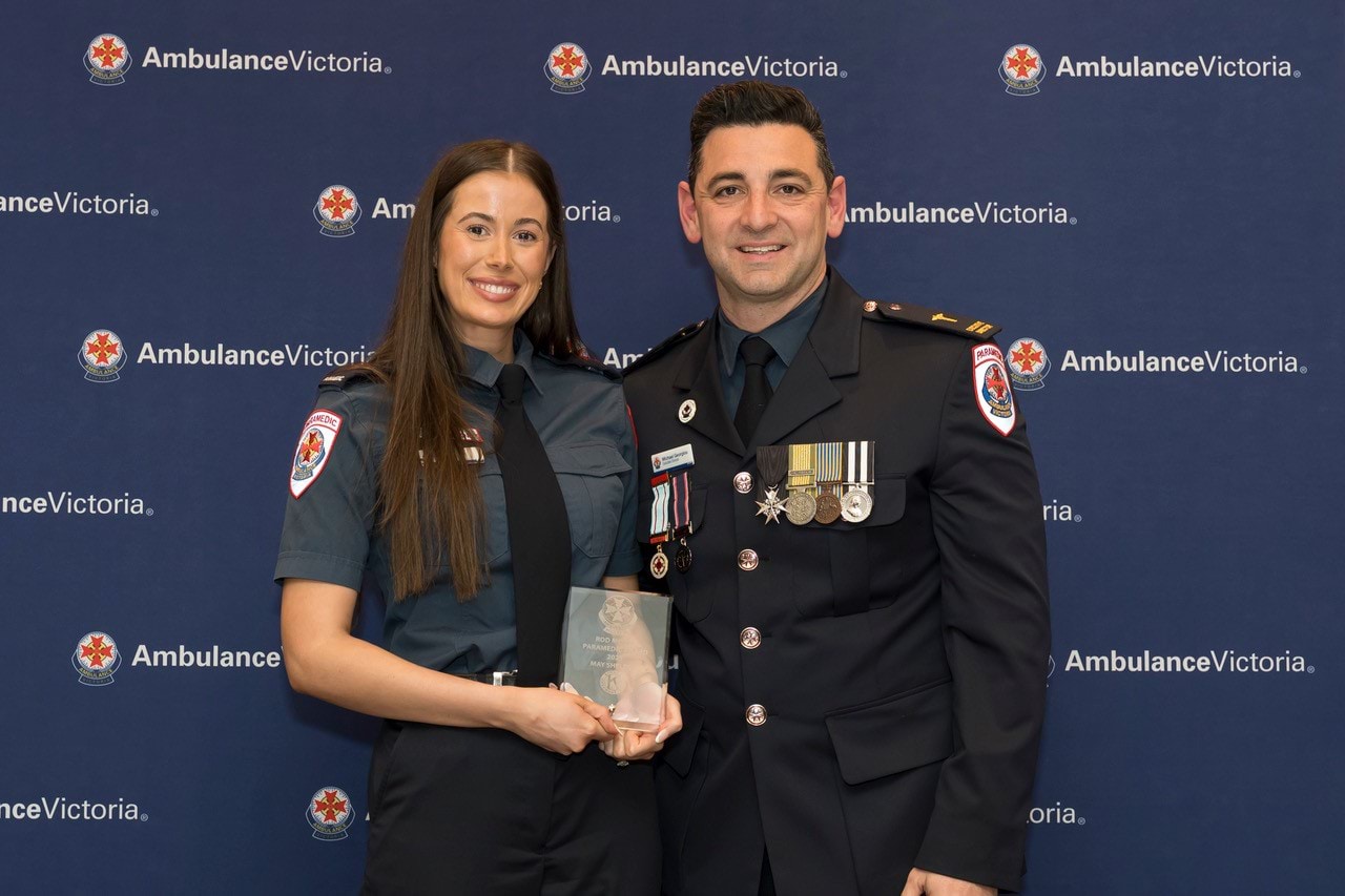 A woman and man in Ambulance Victoria dress uniform smile for the camera. The woman holds an award.